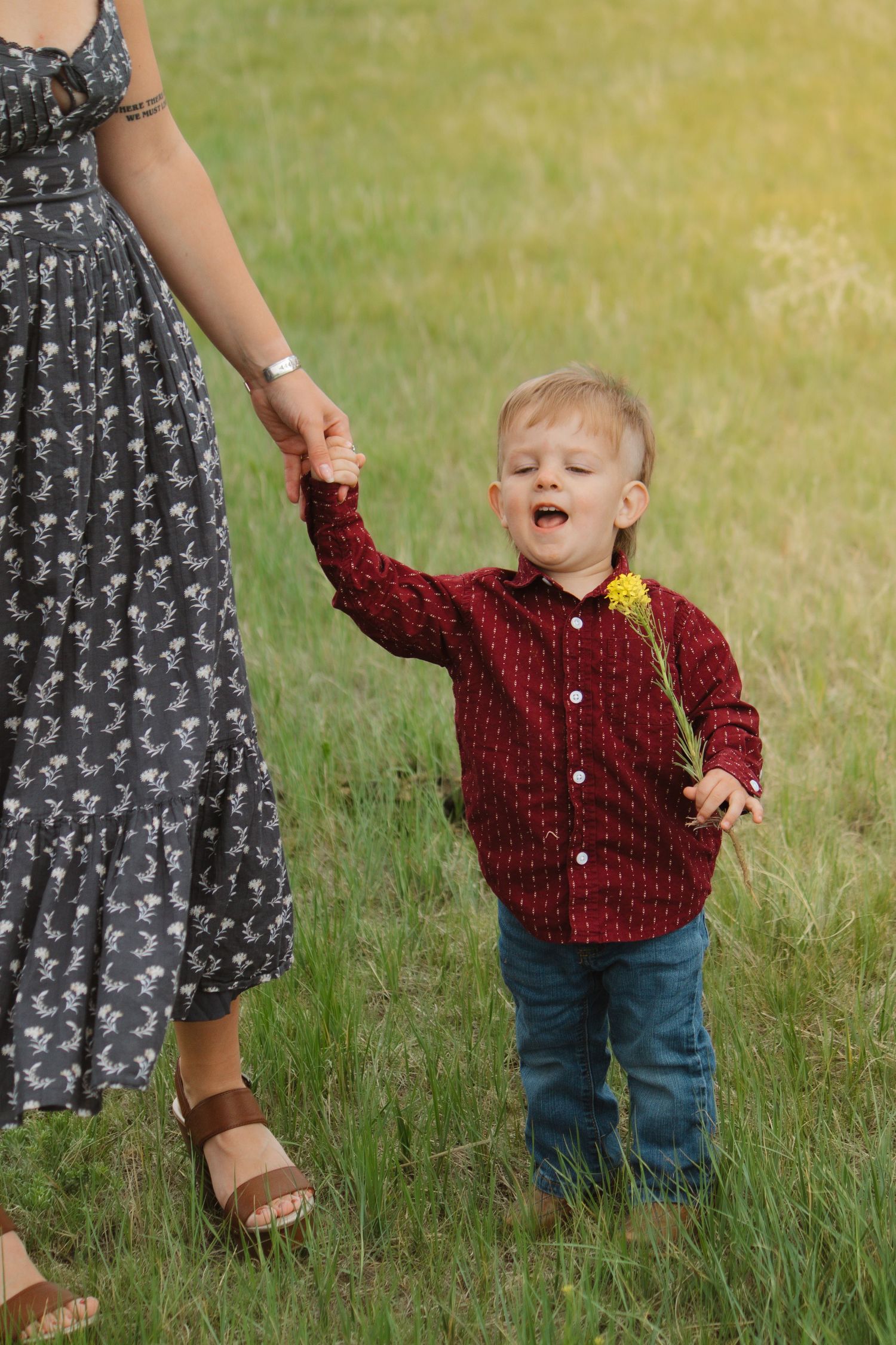 Child in red button-up shirt holds hands with adult in floral dress while walking in grass.