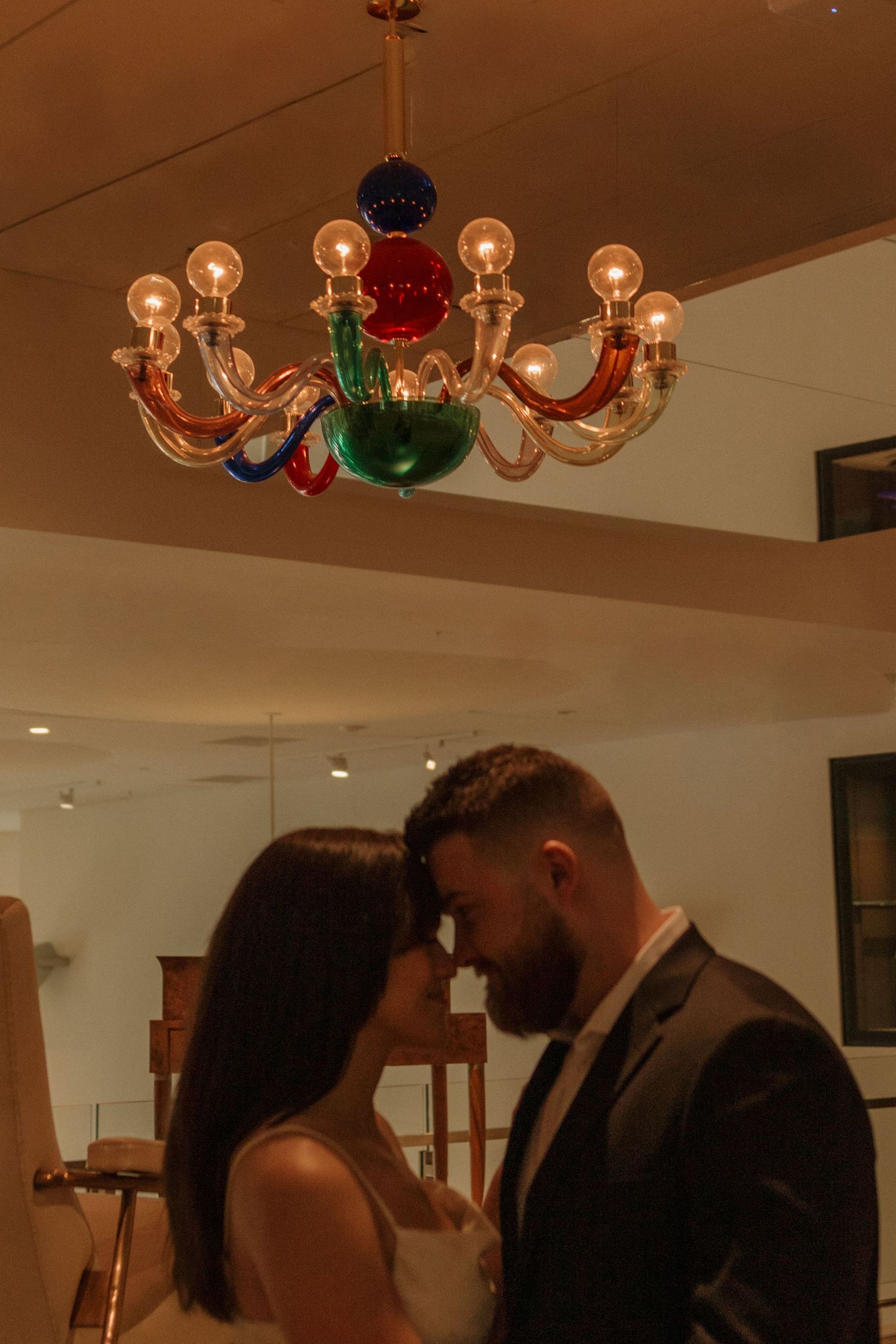 Elegantly dressed couple dancing beneath colorful glass chandeliers in warm lighting.