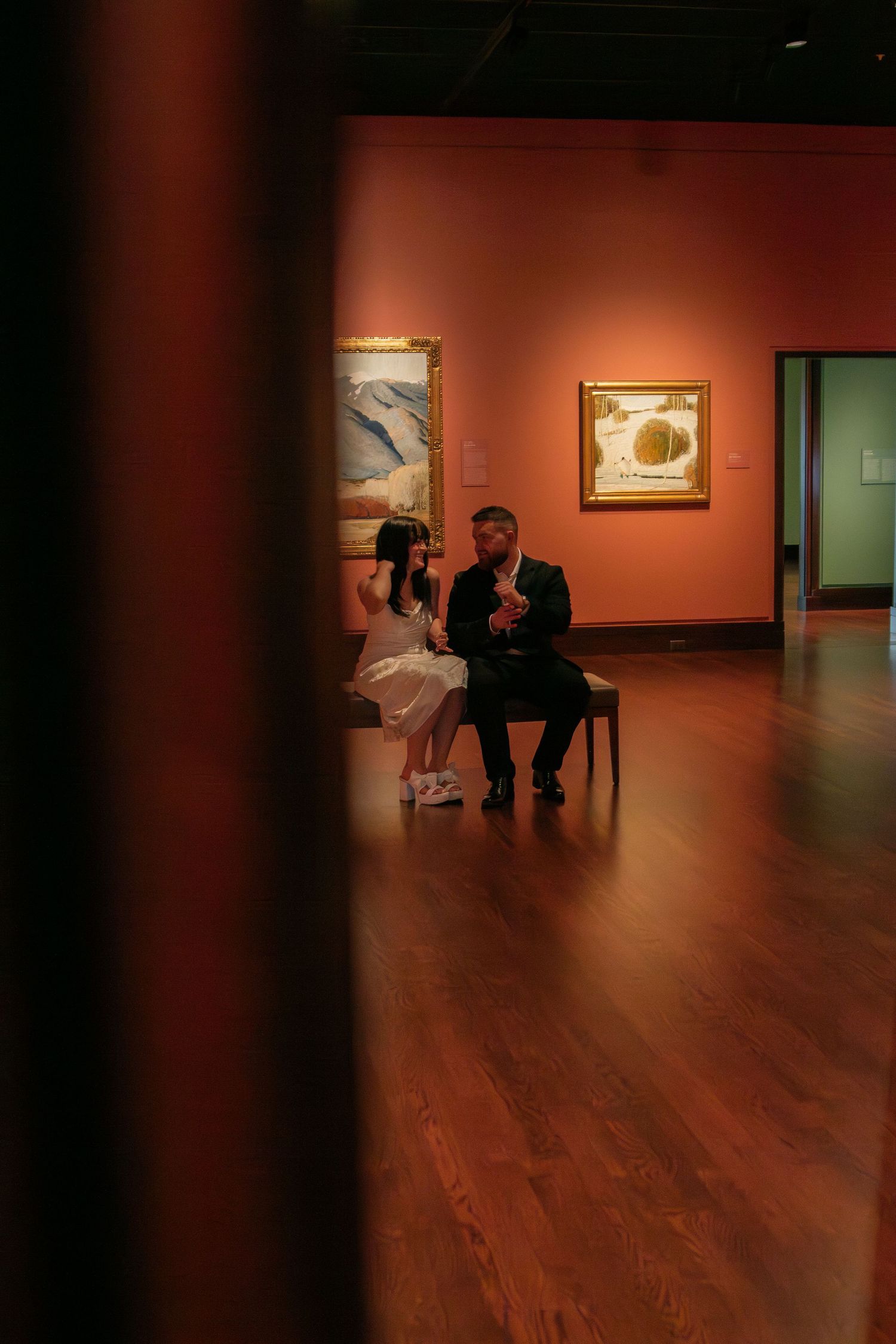 A couple sits together in a dimly lit art gallery with orange walls and framed artwork.