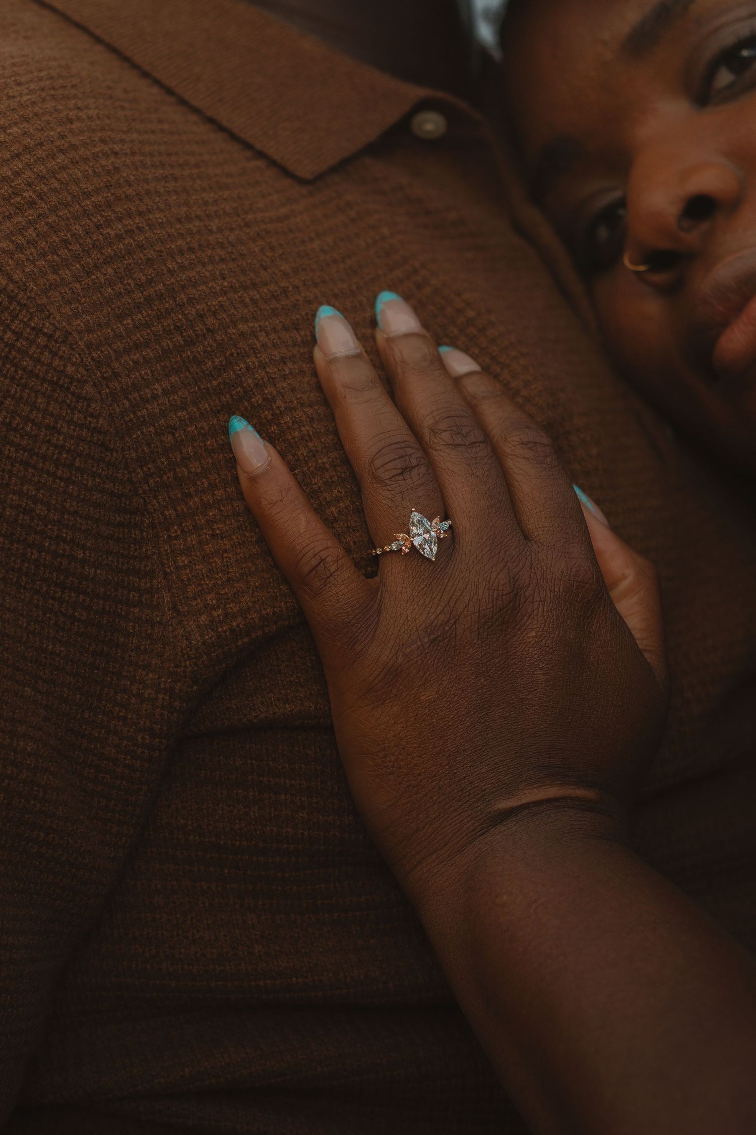 A close-up shot of a hand wearing an engagement ring resting against a brown sweater.