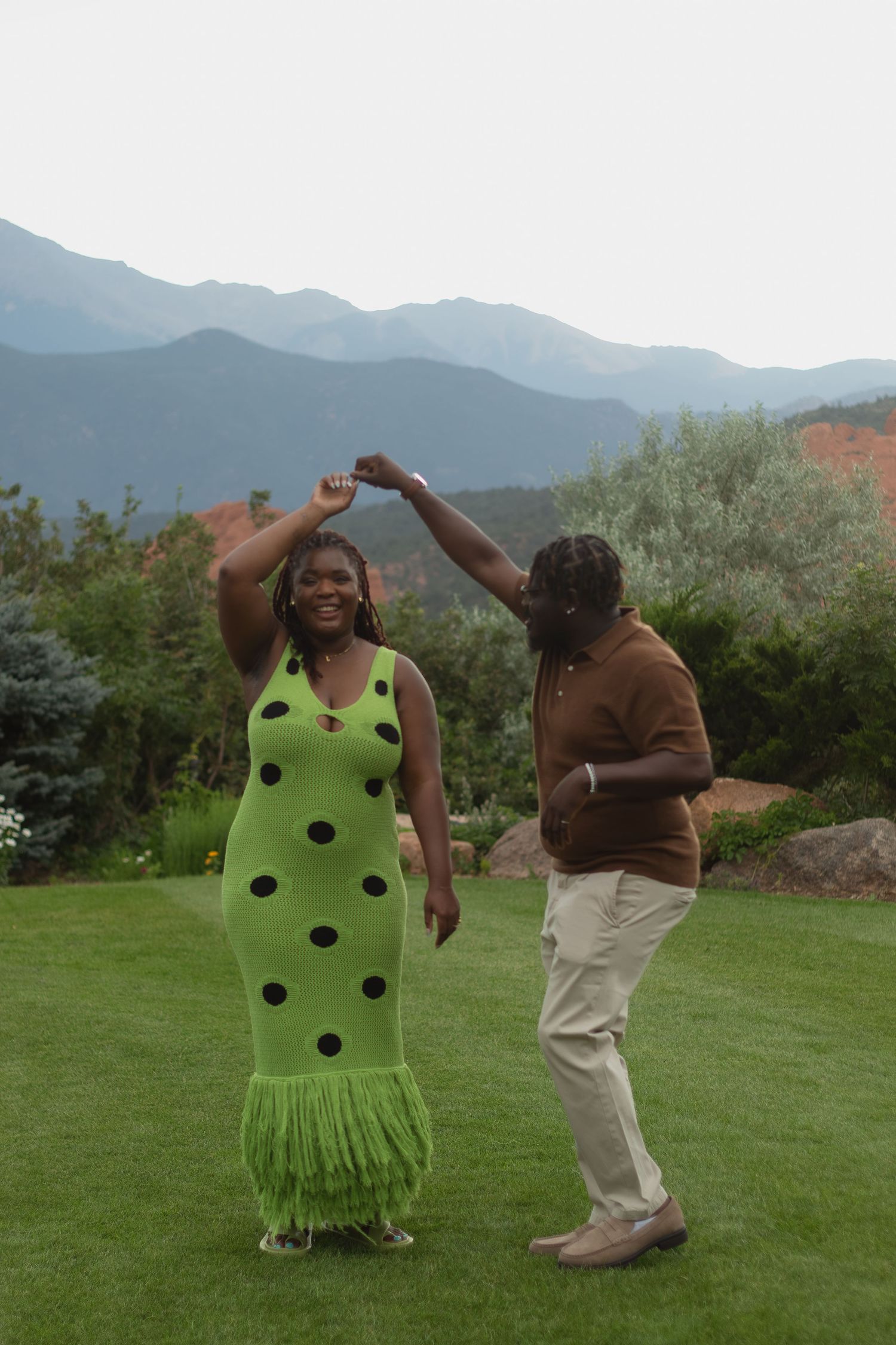 Dancing together on green grass with mountain views, one wearing a bright polka dot dress.