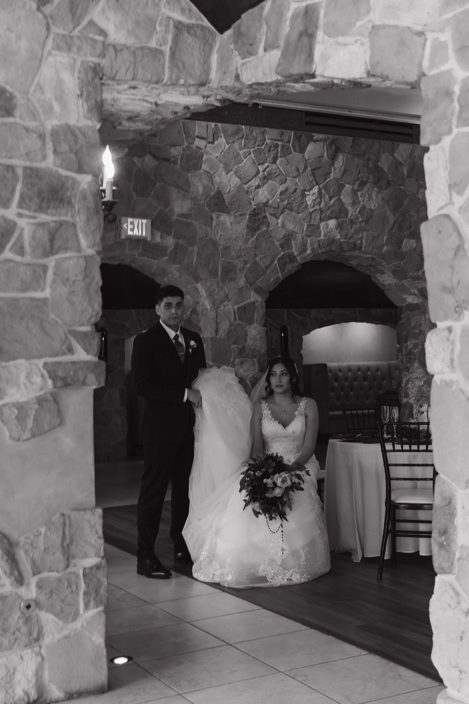 Black and white photo of bride and groom standing in a stone archway holding a floral bouquet.
