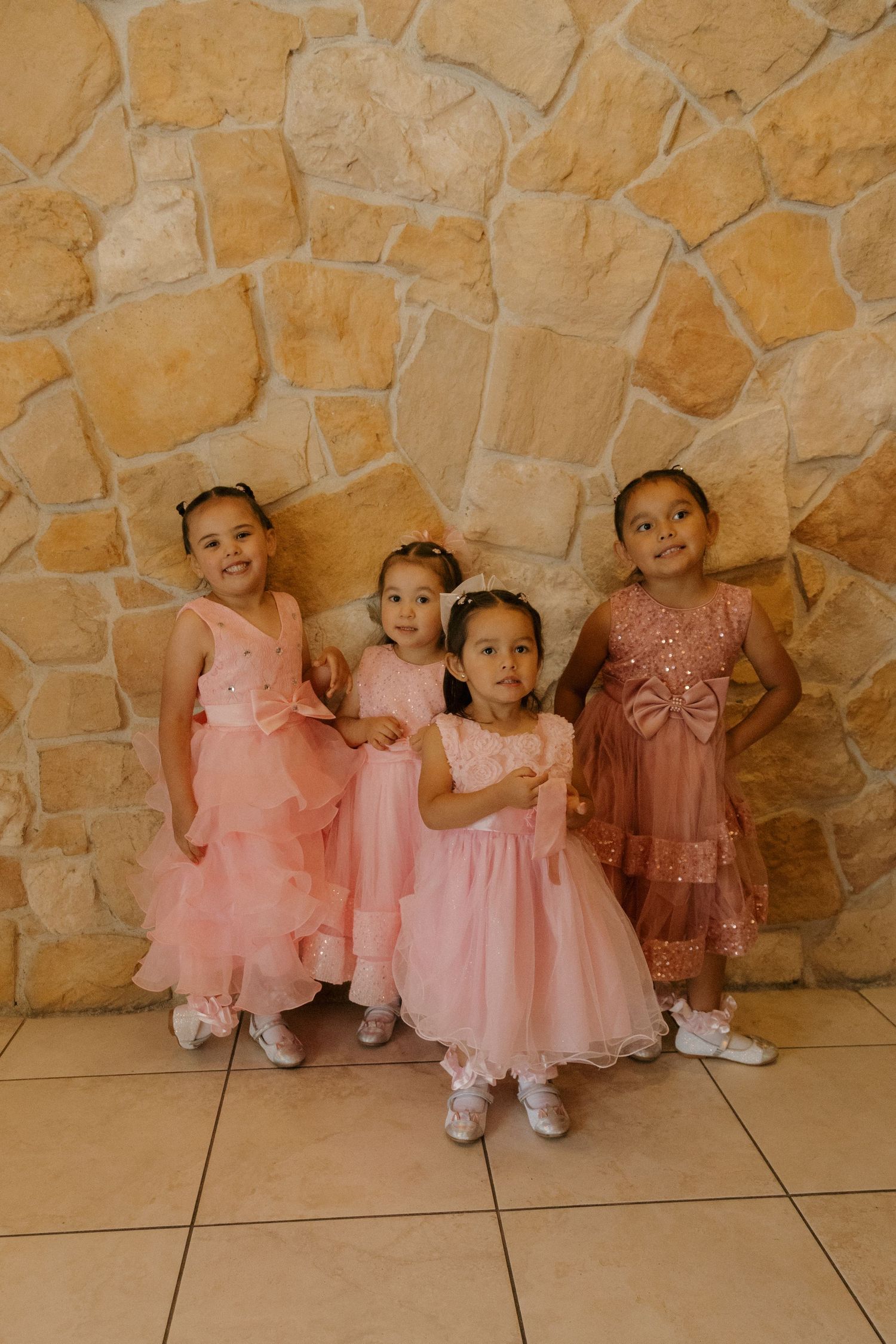 Four young flower girls in pink dresses posing together against a stone wall at a wedding.