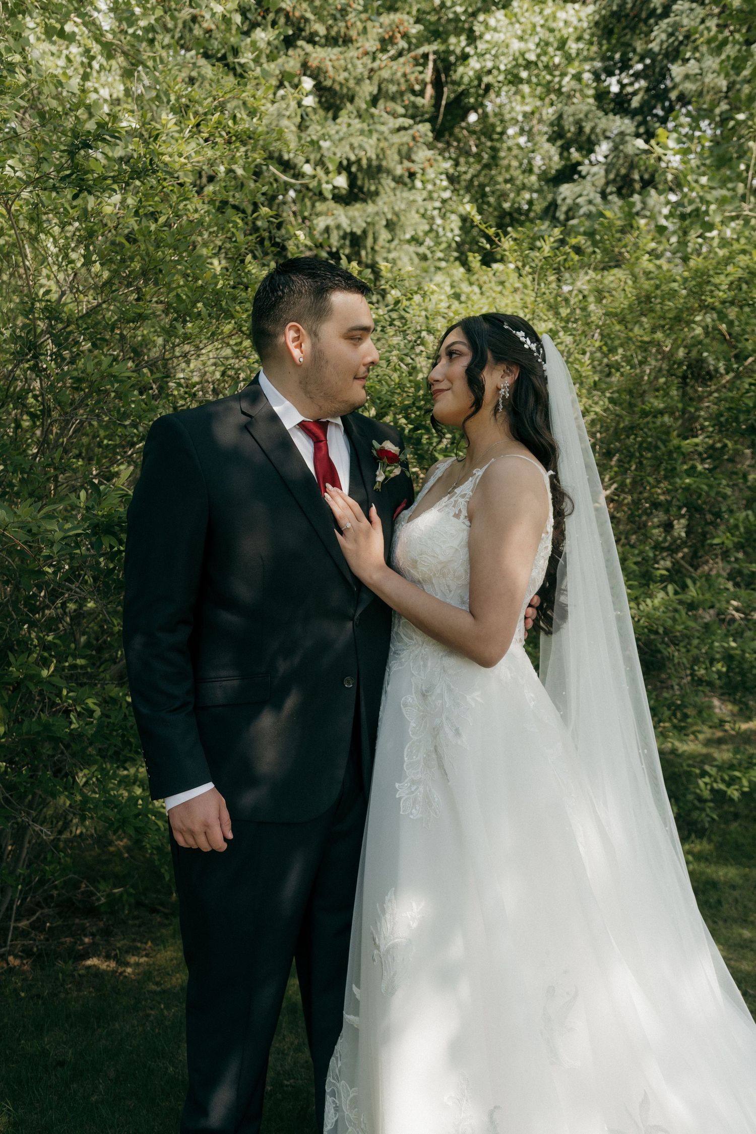 Bride in lace veil and gown gazes at groom in black suit with red tie among green garden trees.