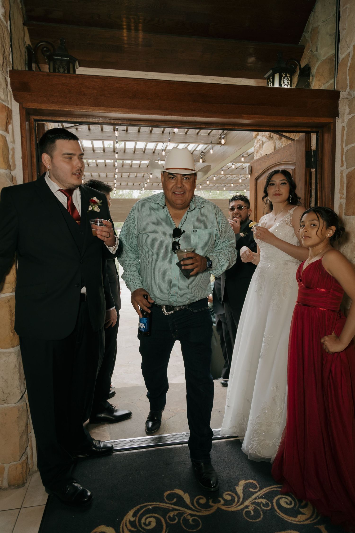Wedding party gathers at venue entrance with bride in white gown and guests in formal attire.