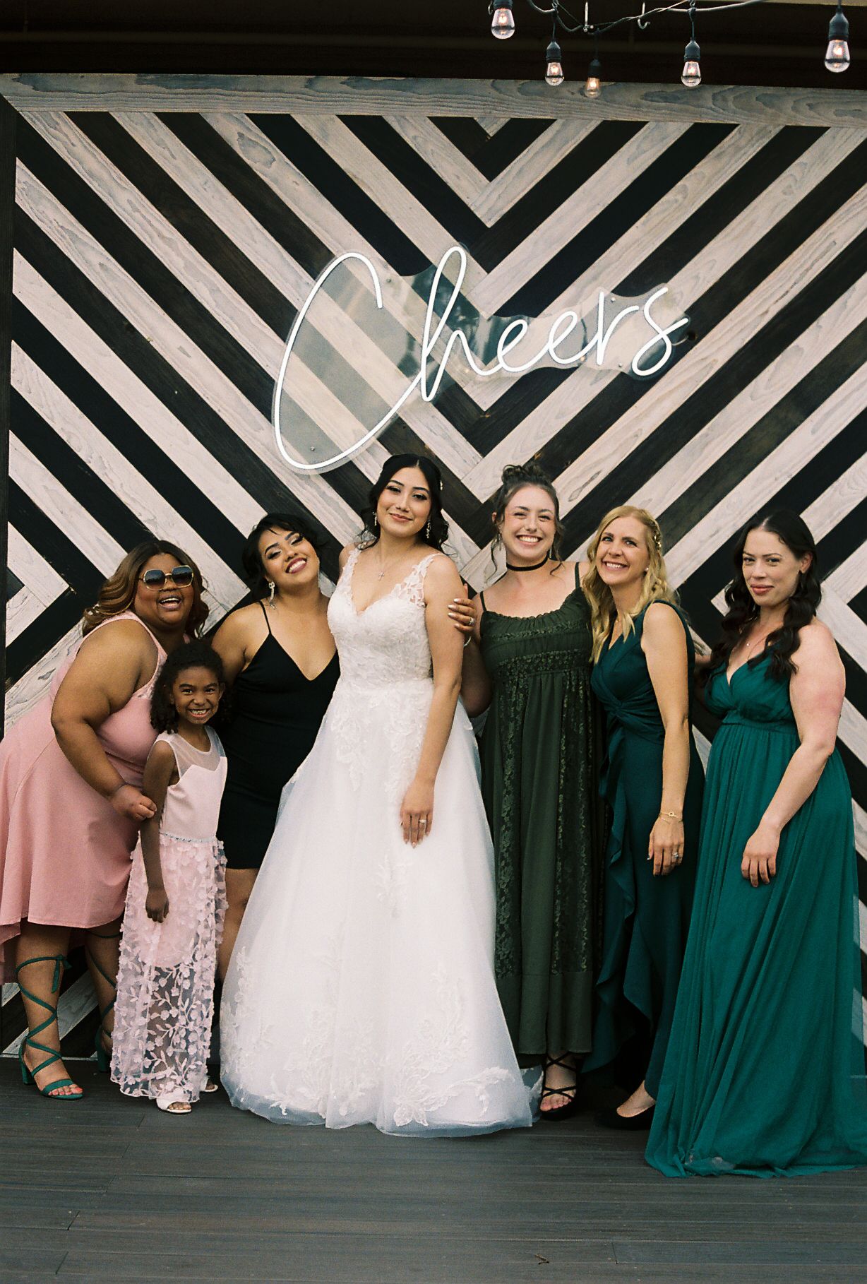 Bride in white gown poses with bridesmaids and friends in front of Cheers neon sign backdrop.