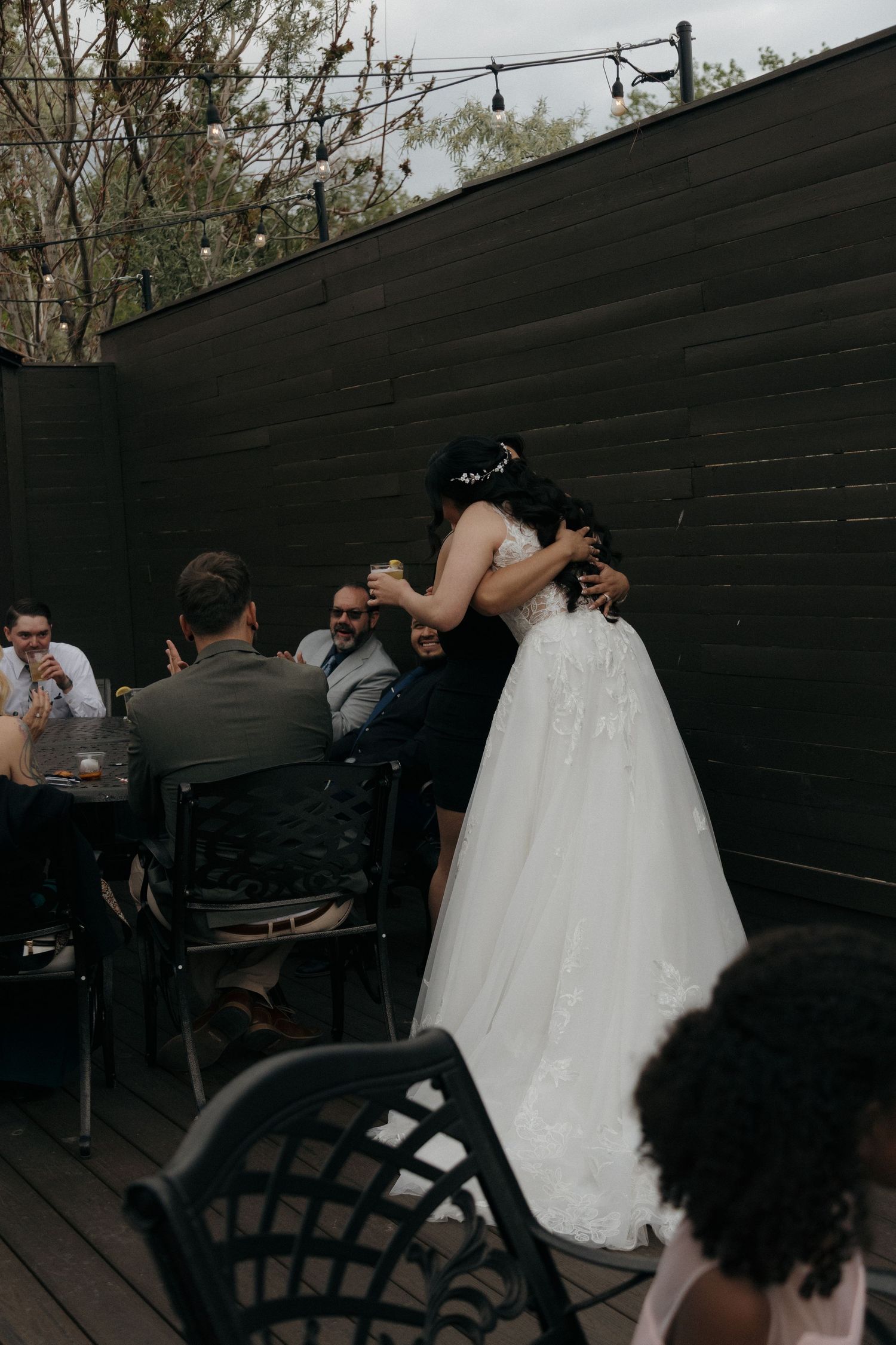 Bride in white gown embraces guest on outdoor patio deck during wedding reception celebration.