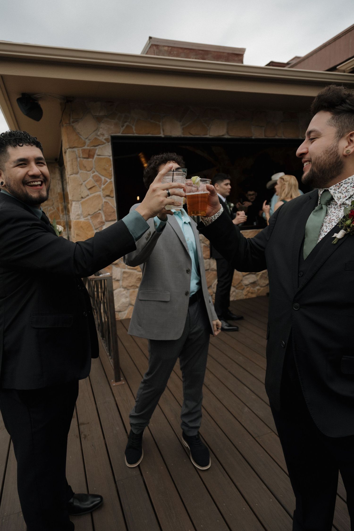 Three men in suits toasting drinks together at an outdoor wedding reception.