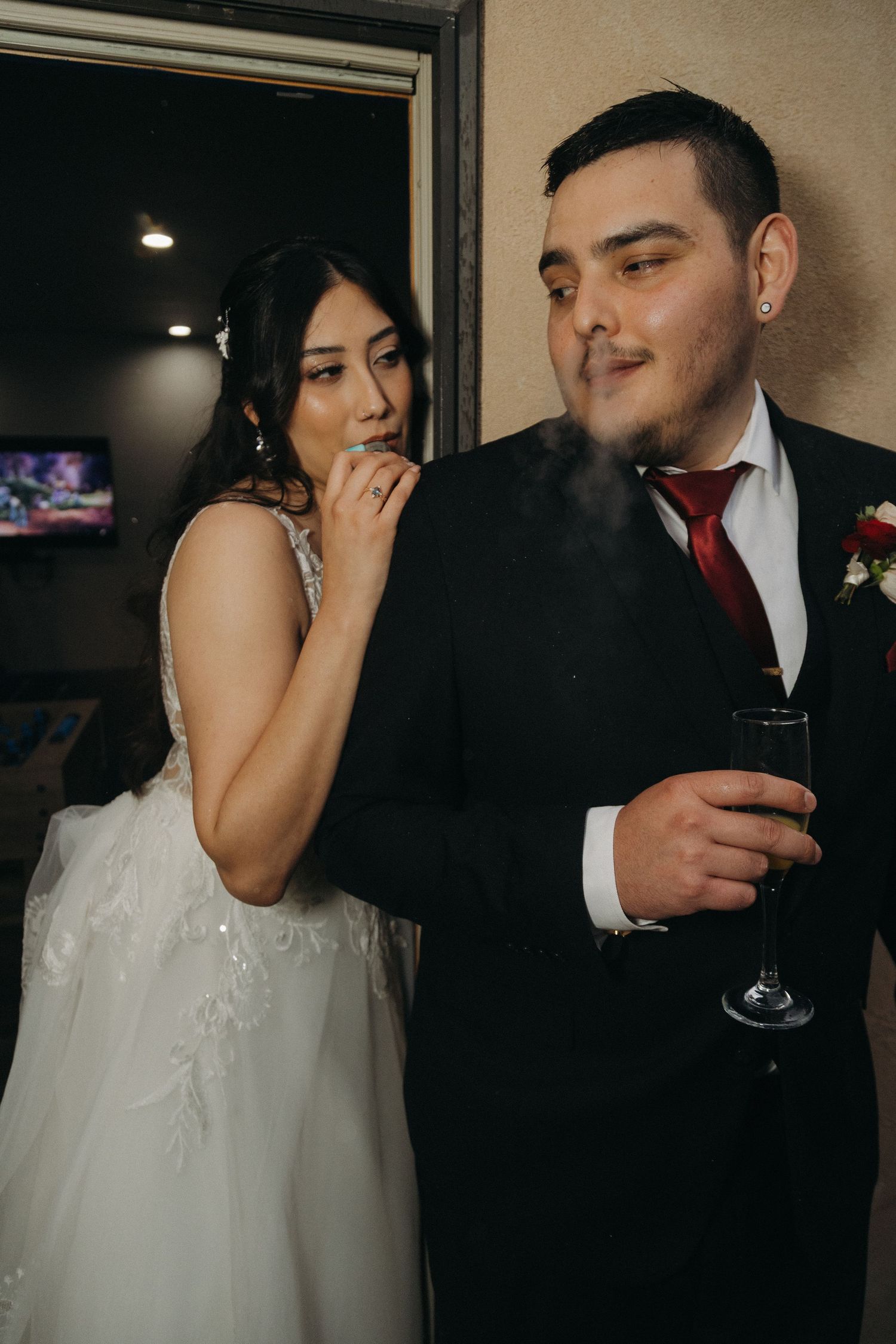 Groom exhaling cigar smoke while bride watches, both holding drinks at their wedding celebration.