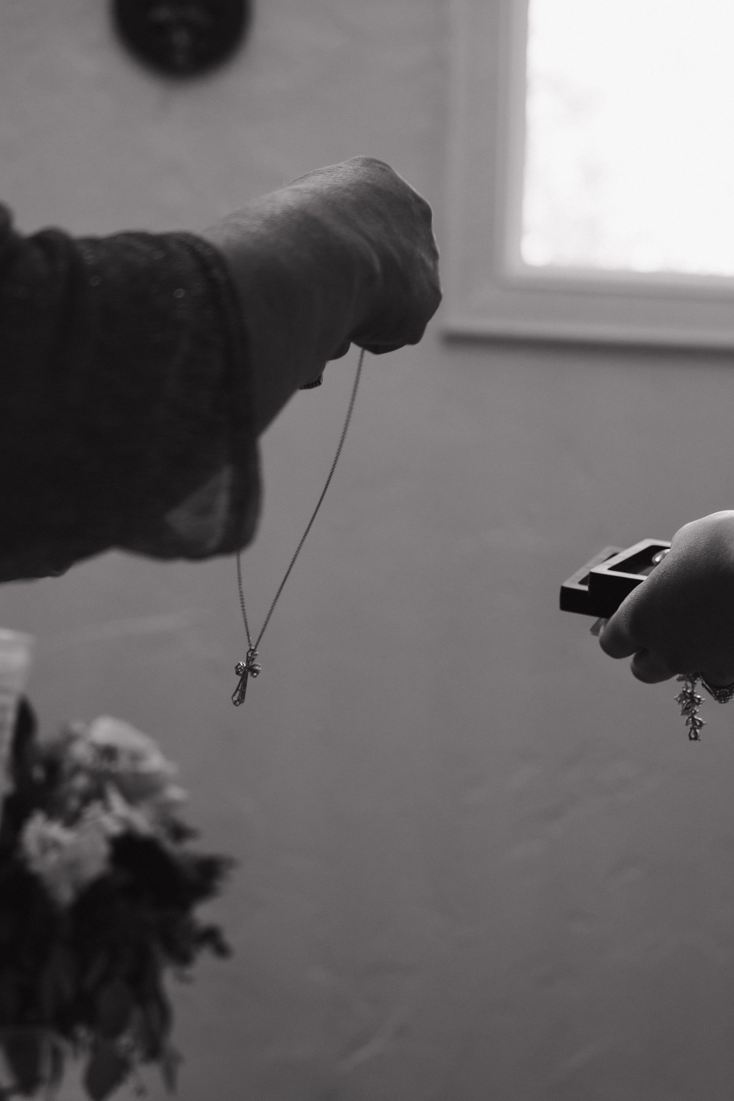 Black and white close-up of hands holding a delicate cross necklace and jewelry box at wedding preparation.