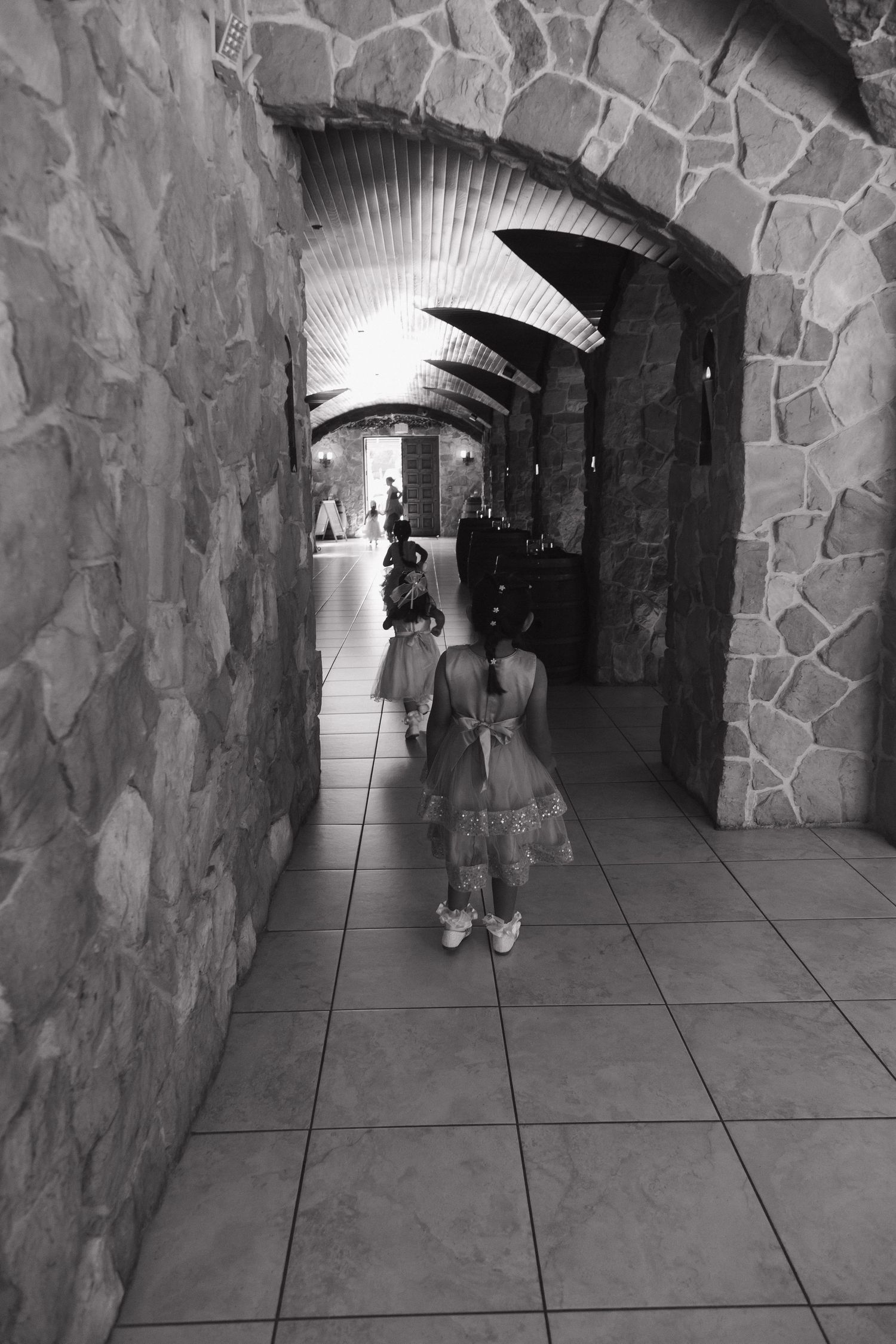 Black and white photo of young flower girls walking through stone archway corridor at wedding venue.