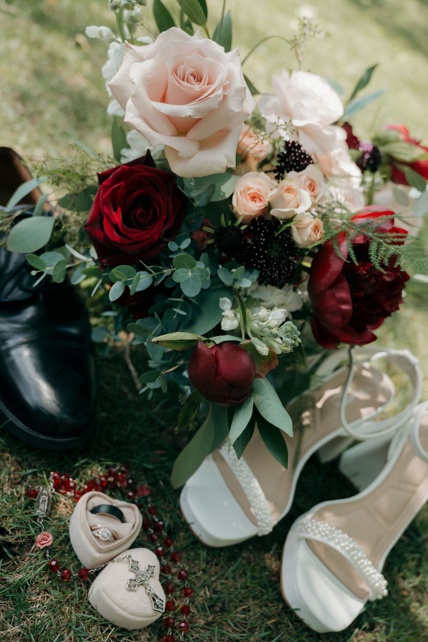 Elegant wedding flat lay with red and blush bouquet, white heels, black dress shoes, and jewelry on green grass.
