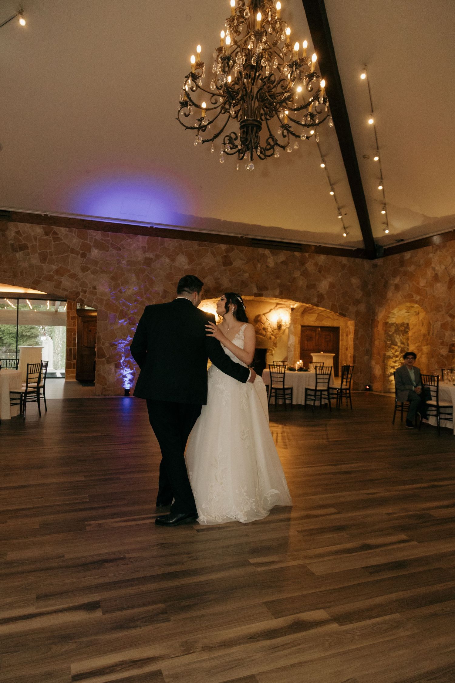 Bride and groom slow dancing under chandelier with purple uplighting in rustic stone wedding venue.