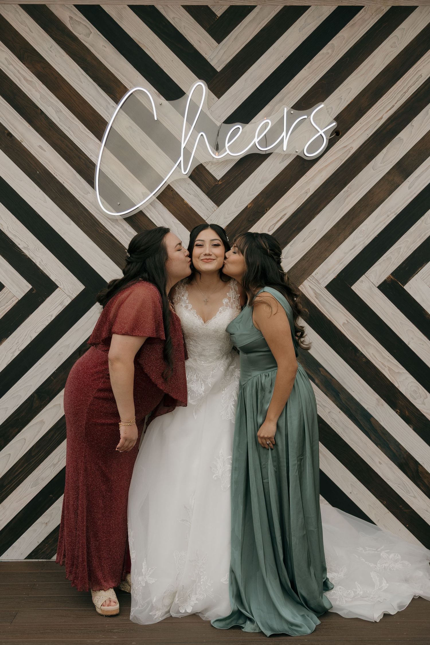 Two guests kiss bride on cheeks in front of a Cheers neon sign at wedding reception photo booth.