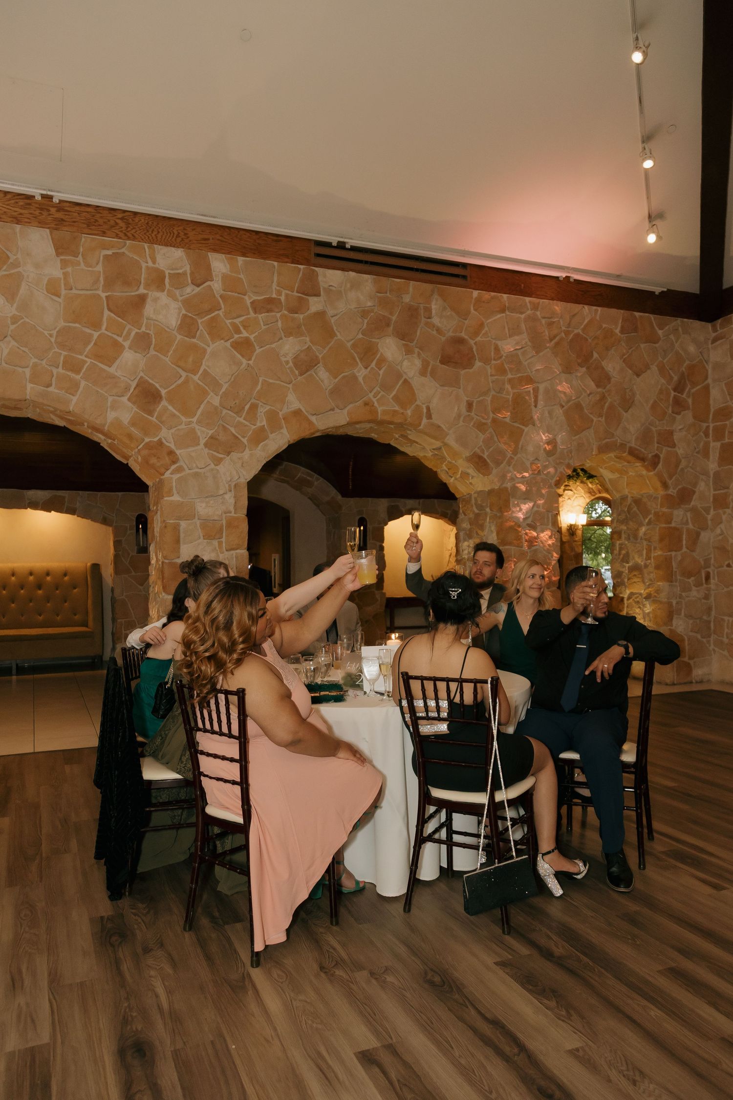 Wedding guests seated at a round table in a rustic stone venue, raising glasses for a celebratory toast.