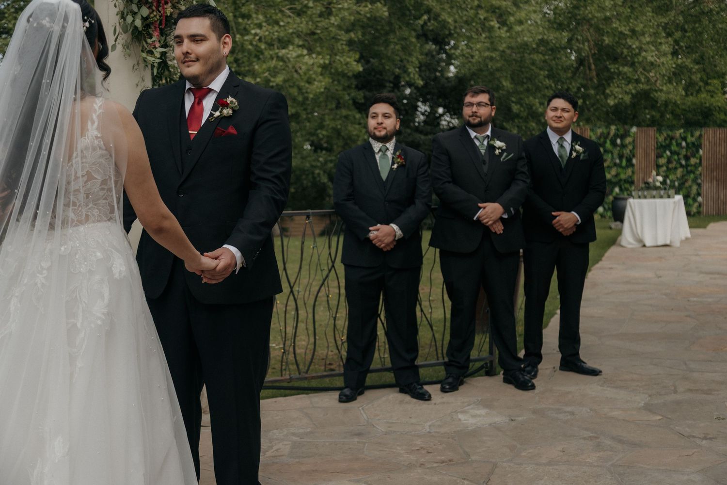A groom and groomsmen in black suits stand at the outdoor wedding altar awaiting the bride.