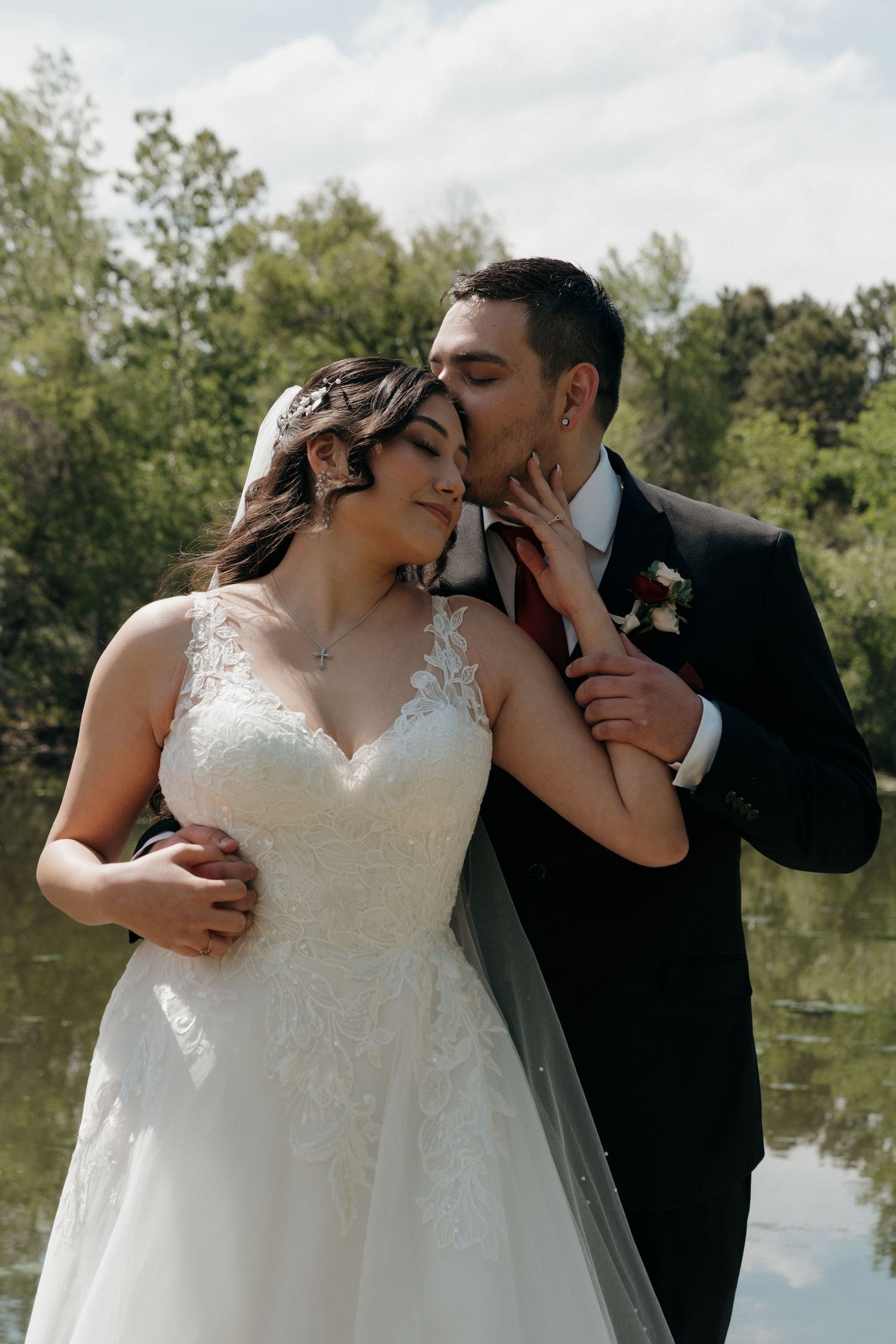 Groom tenderly kisses bride on the cheek near a serene lake backdrop on their wedding day.