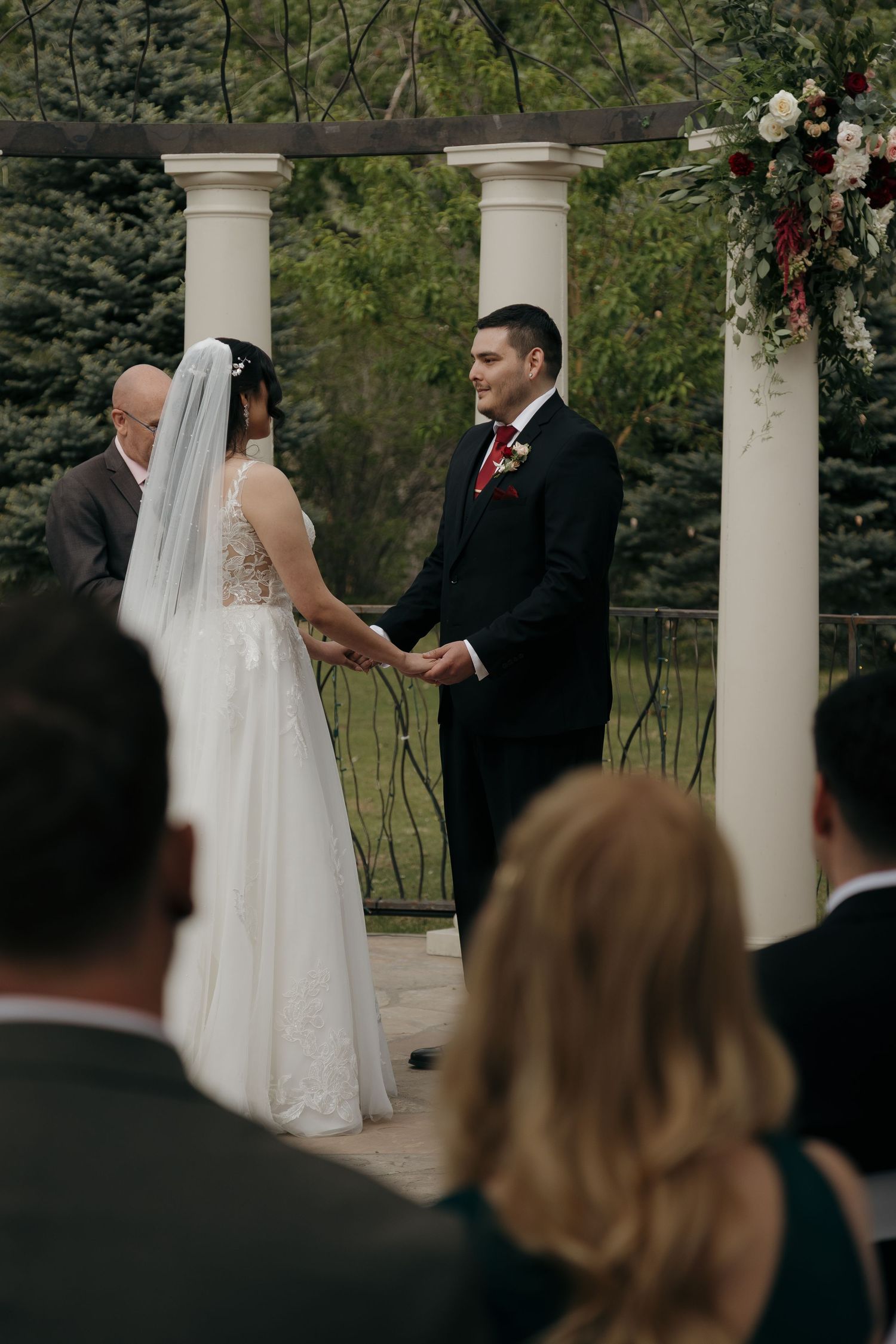 A bride and groom hold hands at an outdoor wedding ceremony beneath a white column pergola.