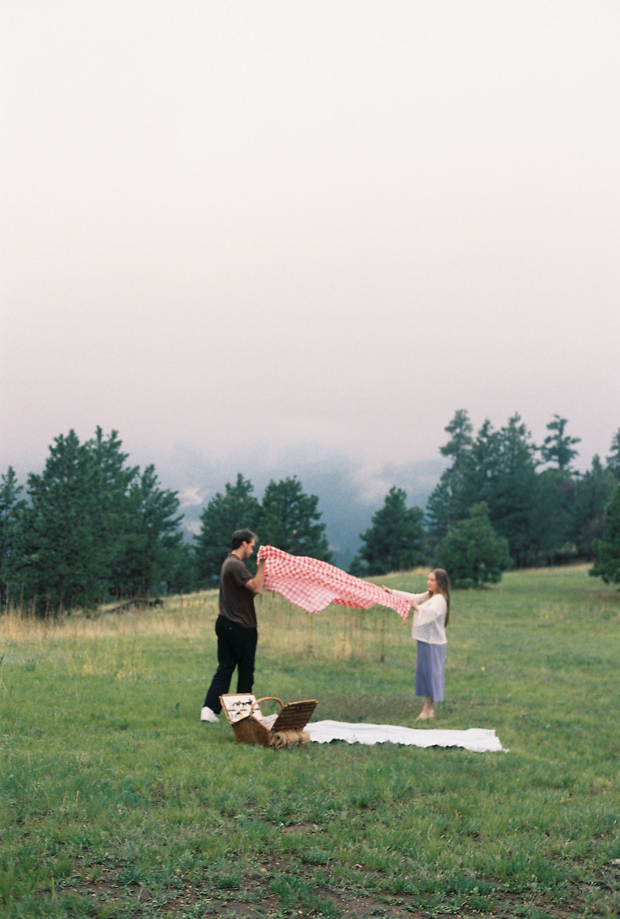 Couple spreading red checkered blanket for picnic in scenic mountain meadow with pine trees and misty sky.