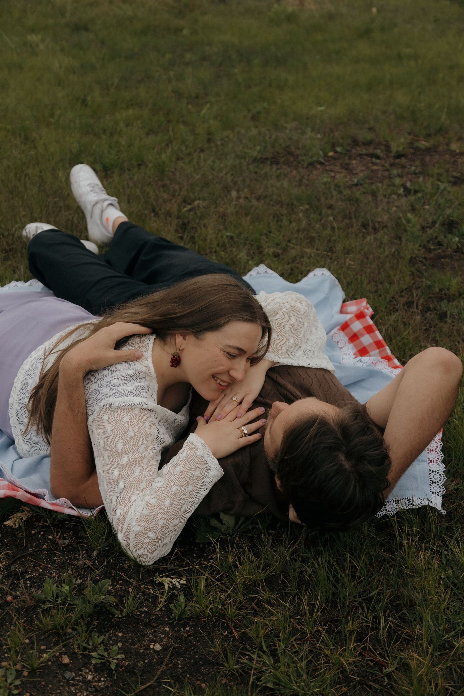 A couple laughs and cuddles together on a blanket in a green grassy field.