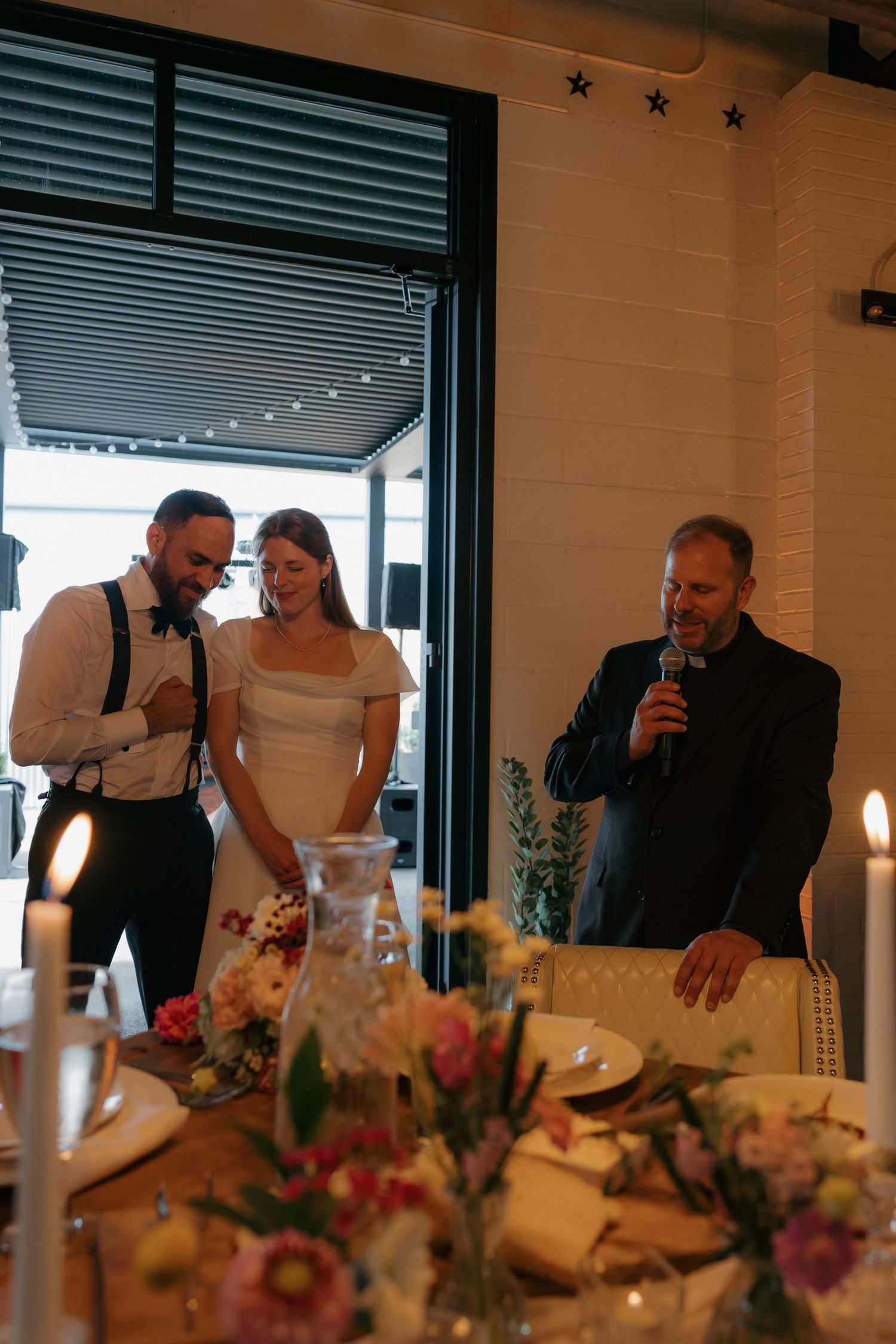 Bride and groom smile as a guest gives a heartfelt speech at an intimate wedding reception dinner.