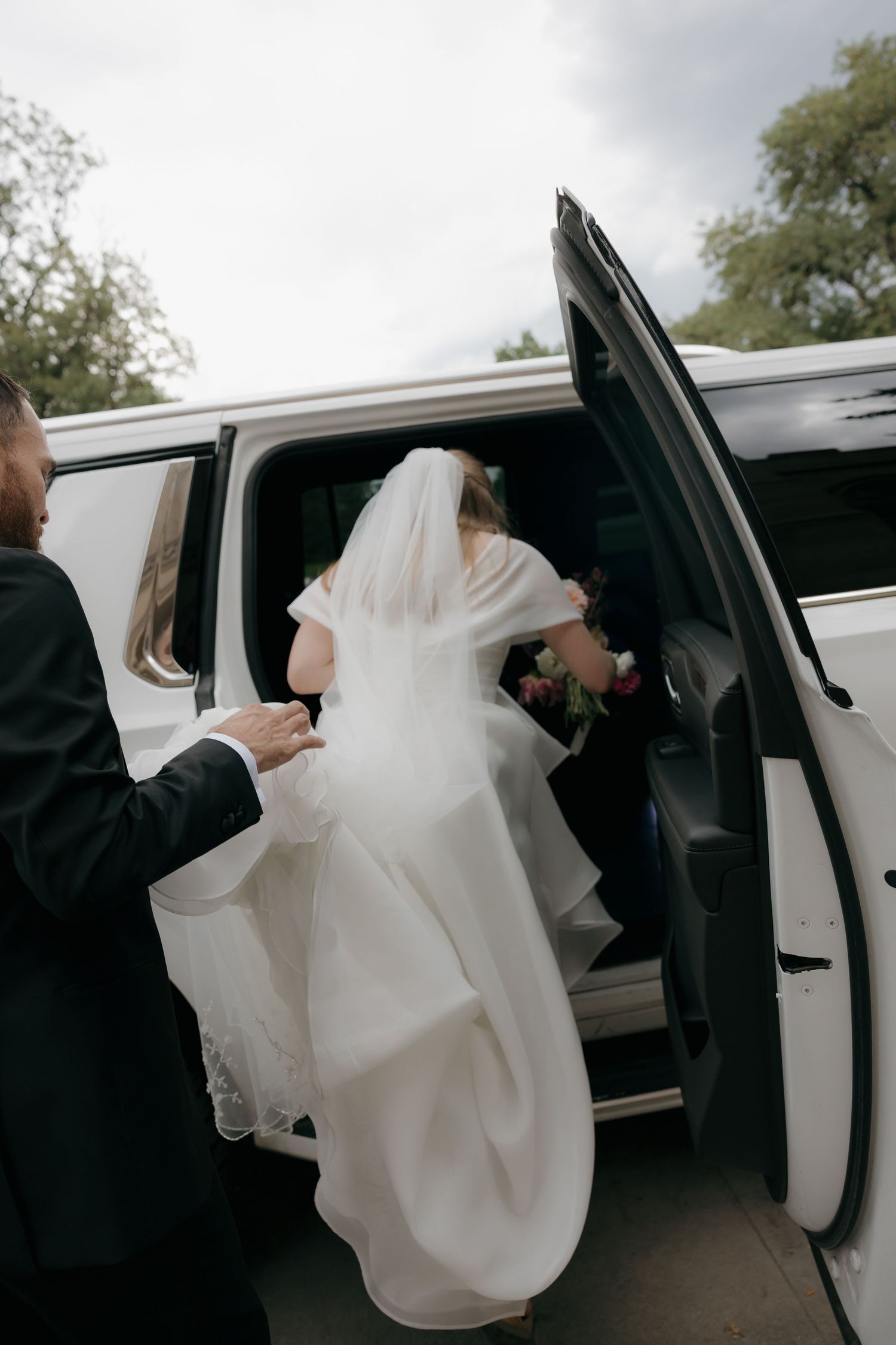 Bride in white gown and veil stepping into a white limousine assisted by groom on wedding day.