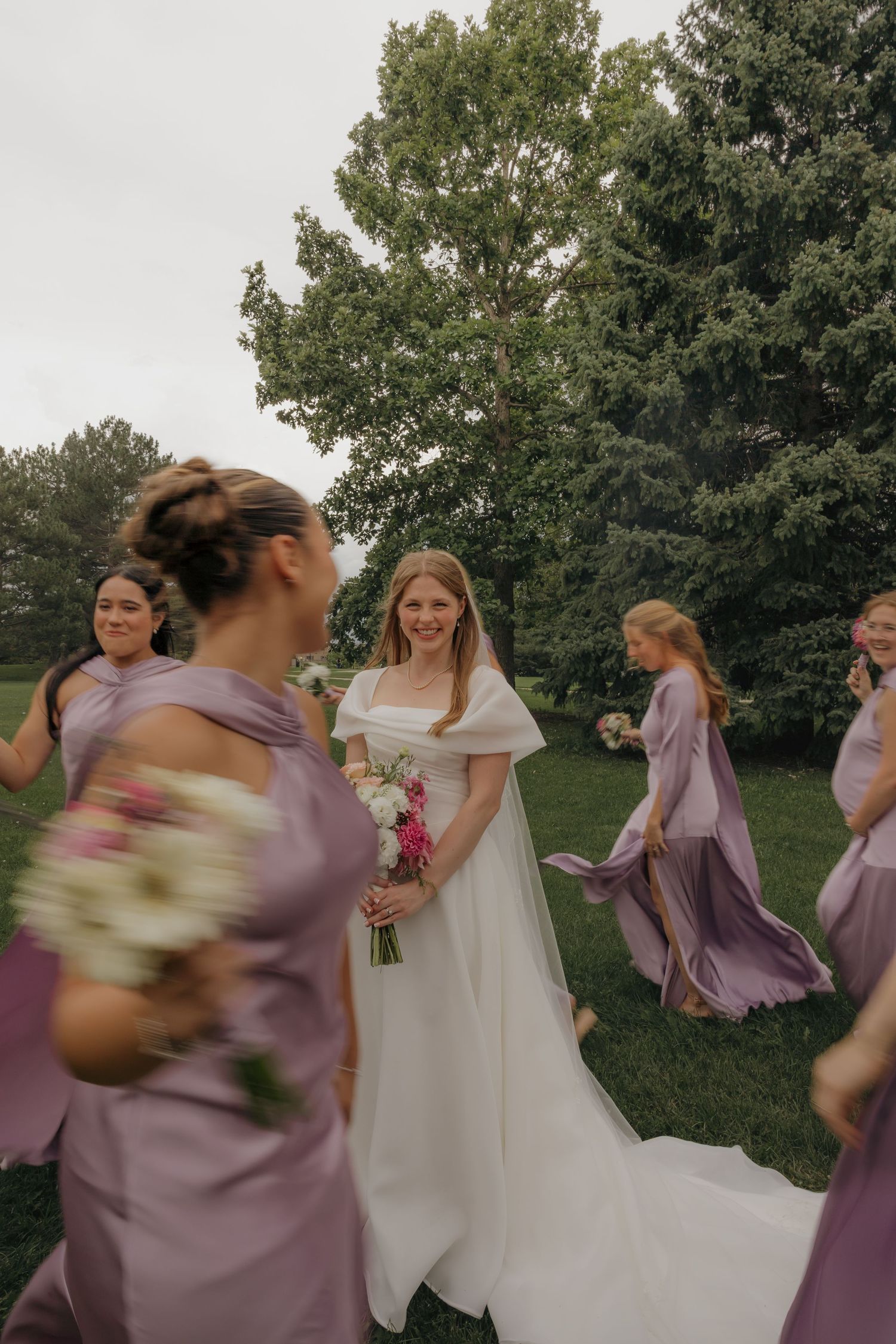 Smiling bride in white off-shoulder gown surrounded by bridesmaids in lavender dresses outdoors.