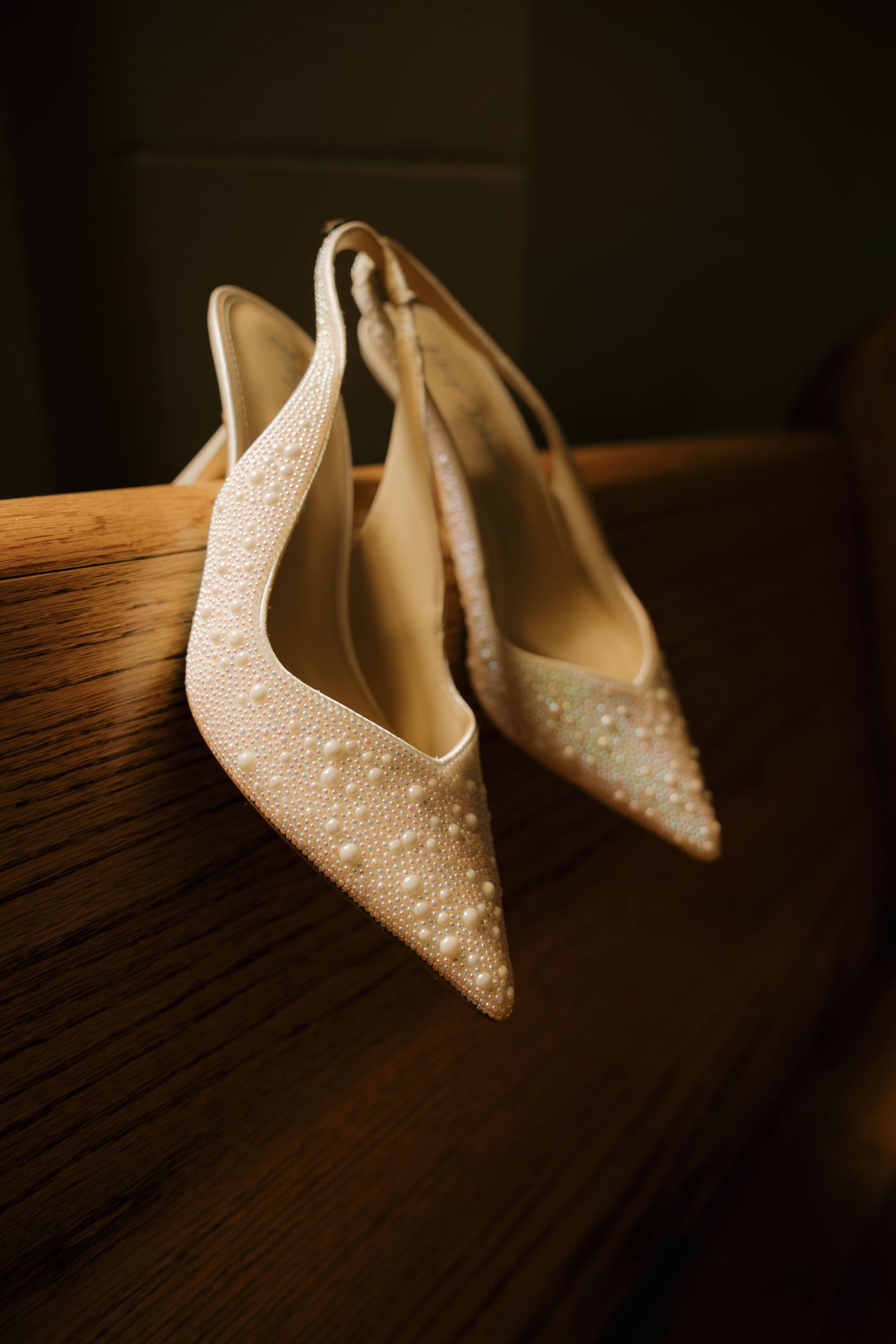 Pearl-embellished ivory slingback wedding heels with pointed toes resting on wooden church pew.