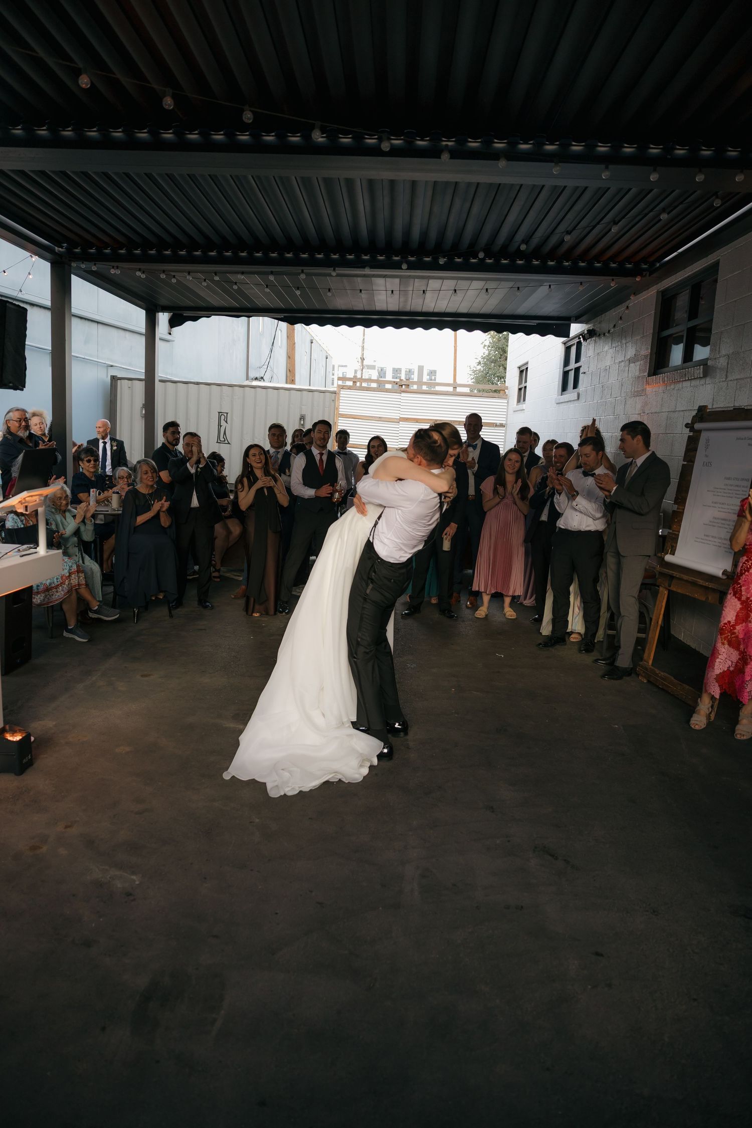 Groom lifts bride in white gown during first dance while wedding guests cheer and applaud in outdoor venue.