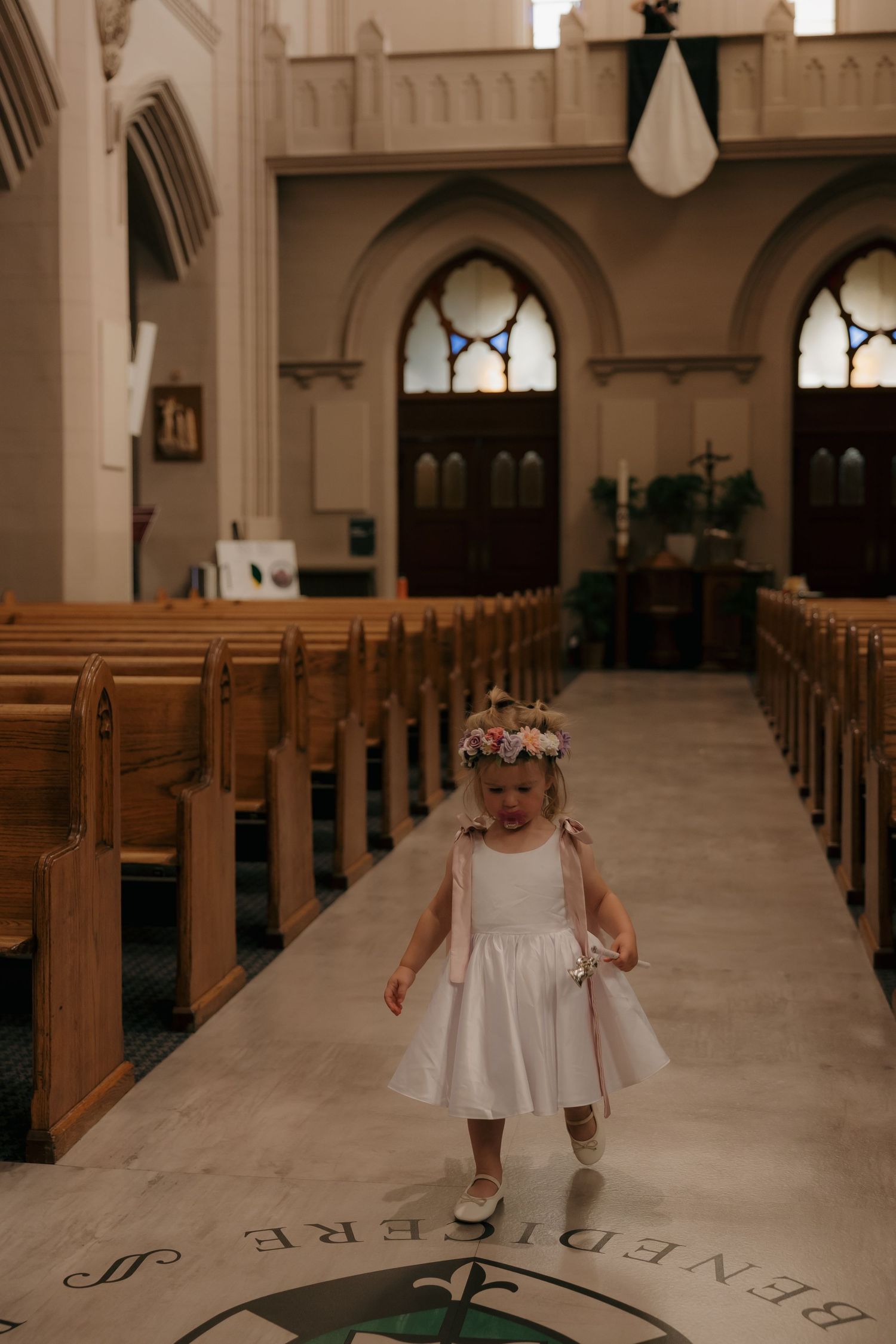 Flower girl in white dress walks down Catholic church aisle wearing floral crown.