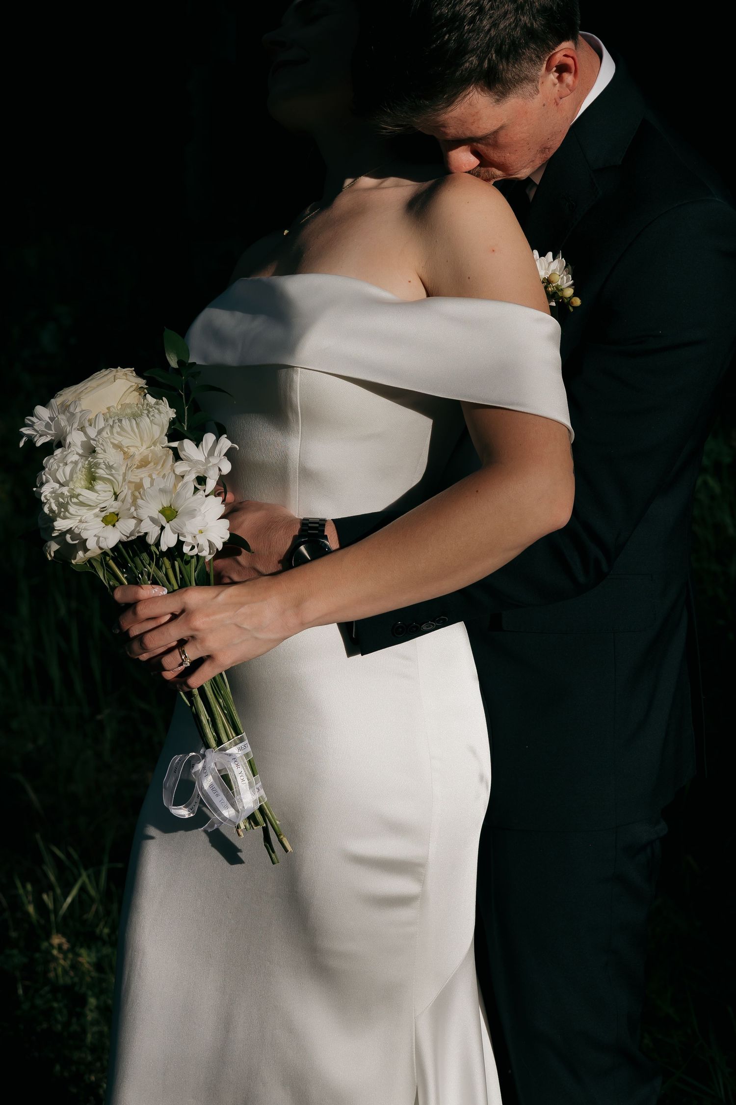 Bride in off-shoulder white gown embraces groom in black suit while holding white floral bouquet.