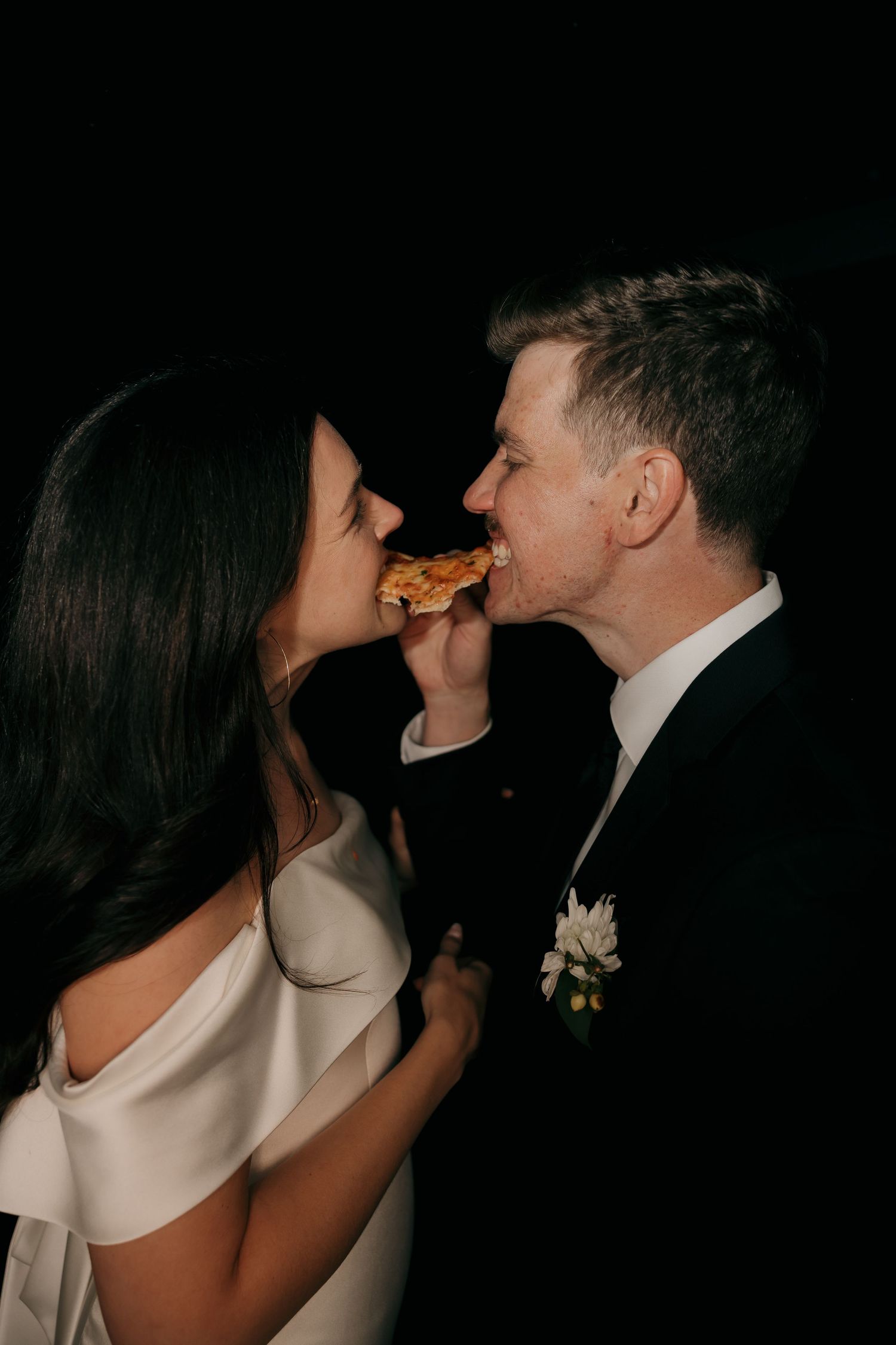Bride and groom sharing a slice of pizza together at their wedding reception against a dark background.