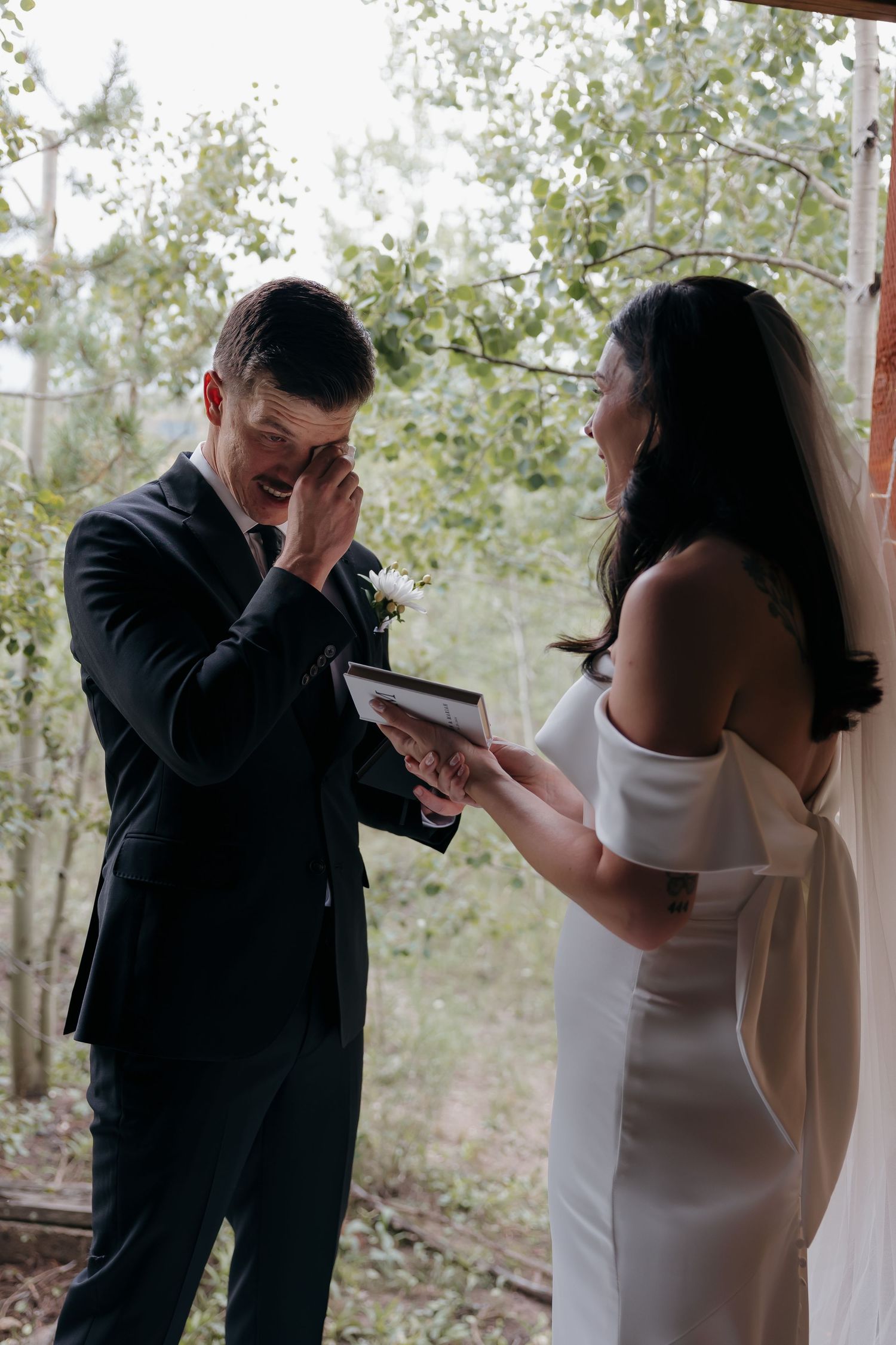 Emotional groom wiping tears as bride reads heartfelt vows during outdoor ceremony.