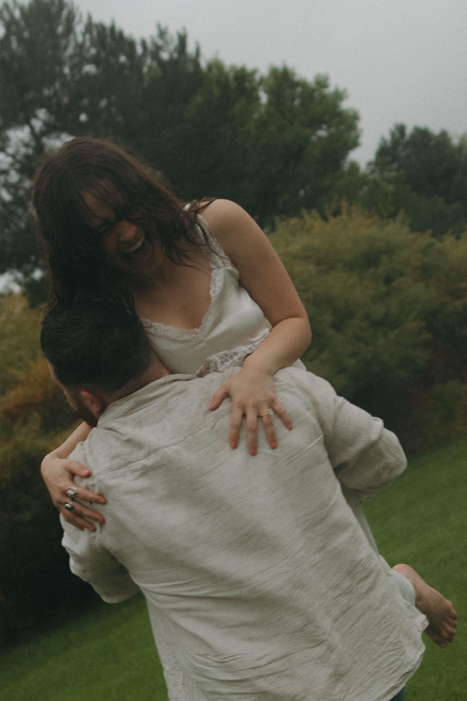 Person in a white dress spins playfully in a grassy outdoor setting surrounded by trees.