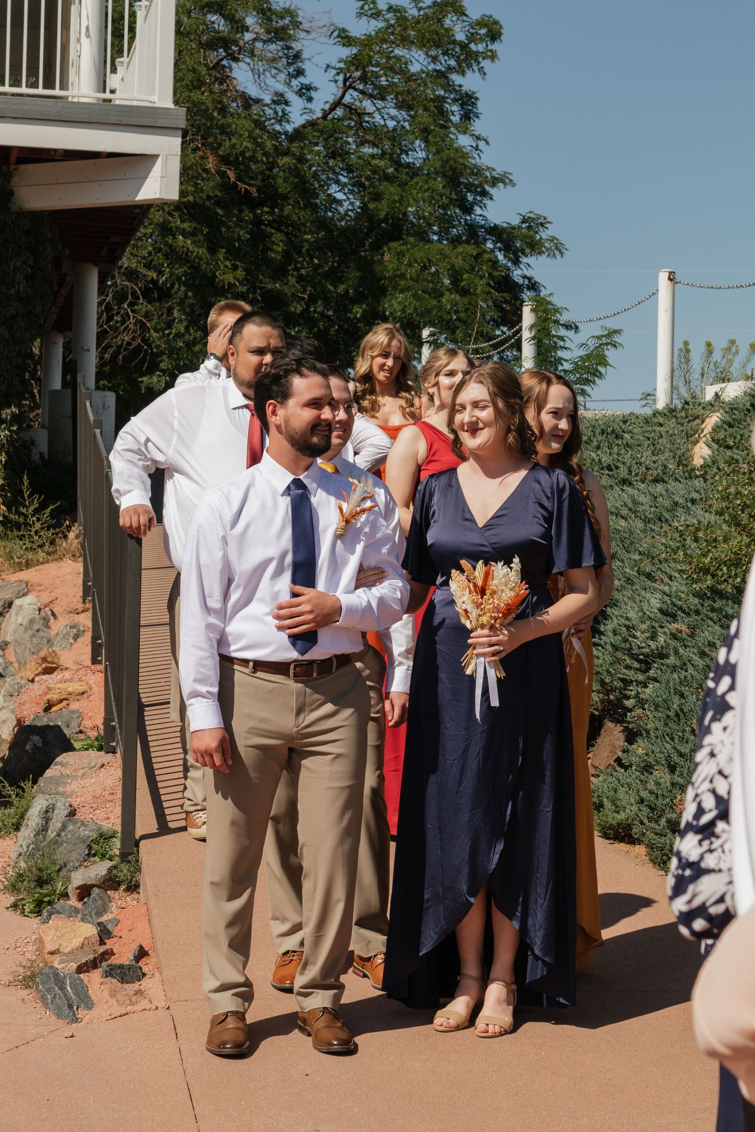 A wedding party stands together on an outdoor patio surrounded by greenery on a sunny day.
