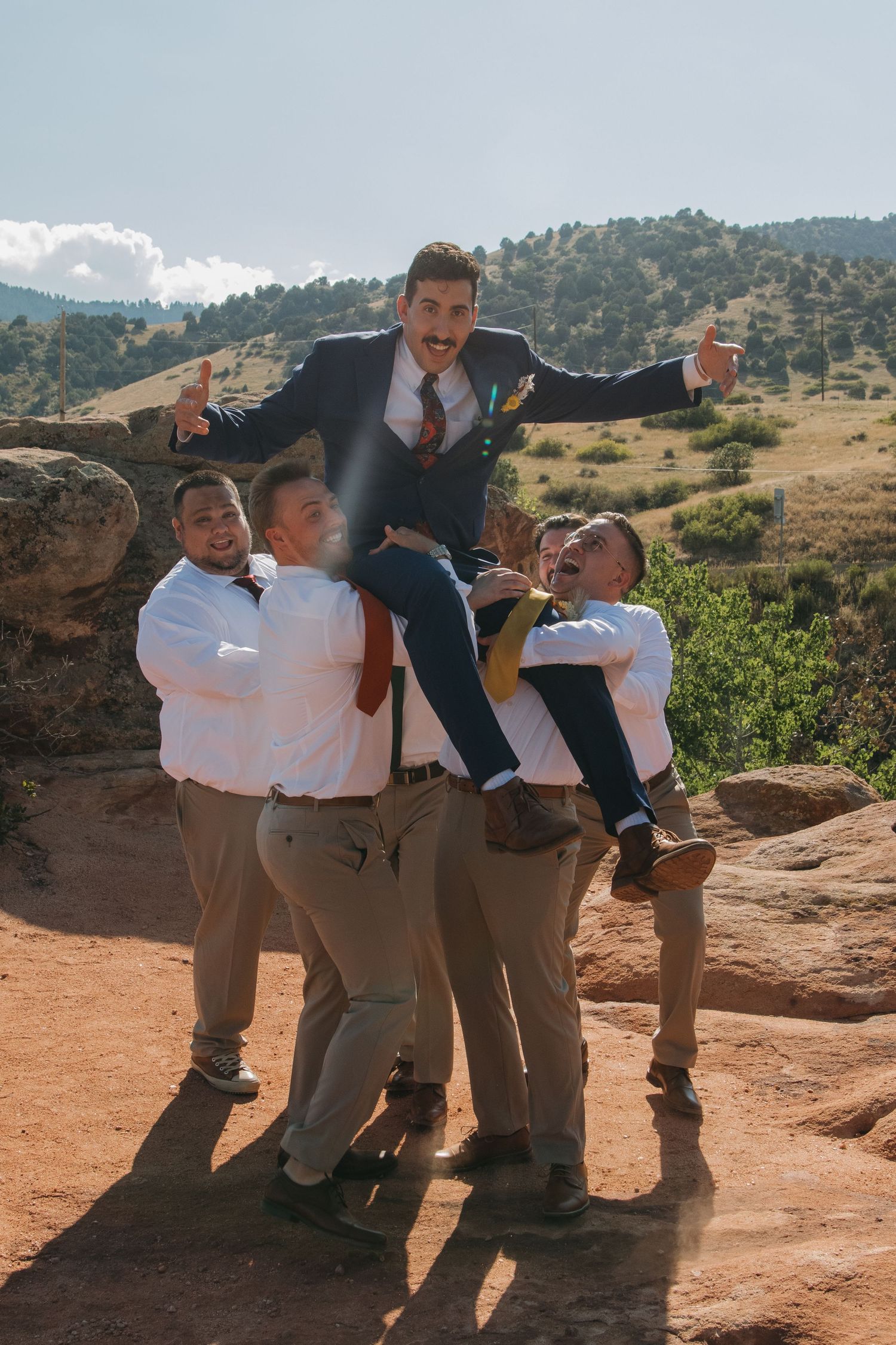 Groomsmen lift and support someone in a playful pose on red rocks during wedding festivities.
