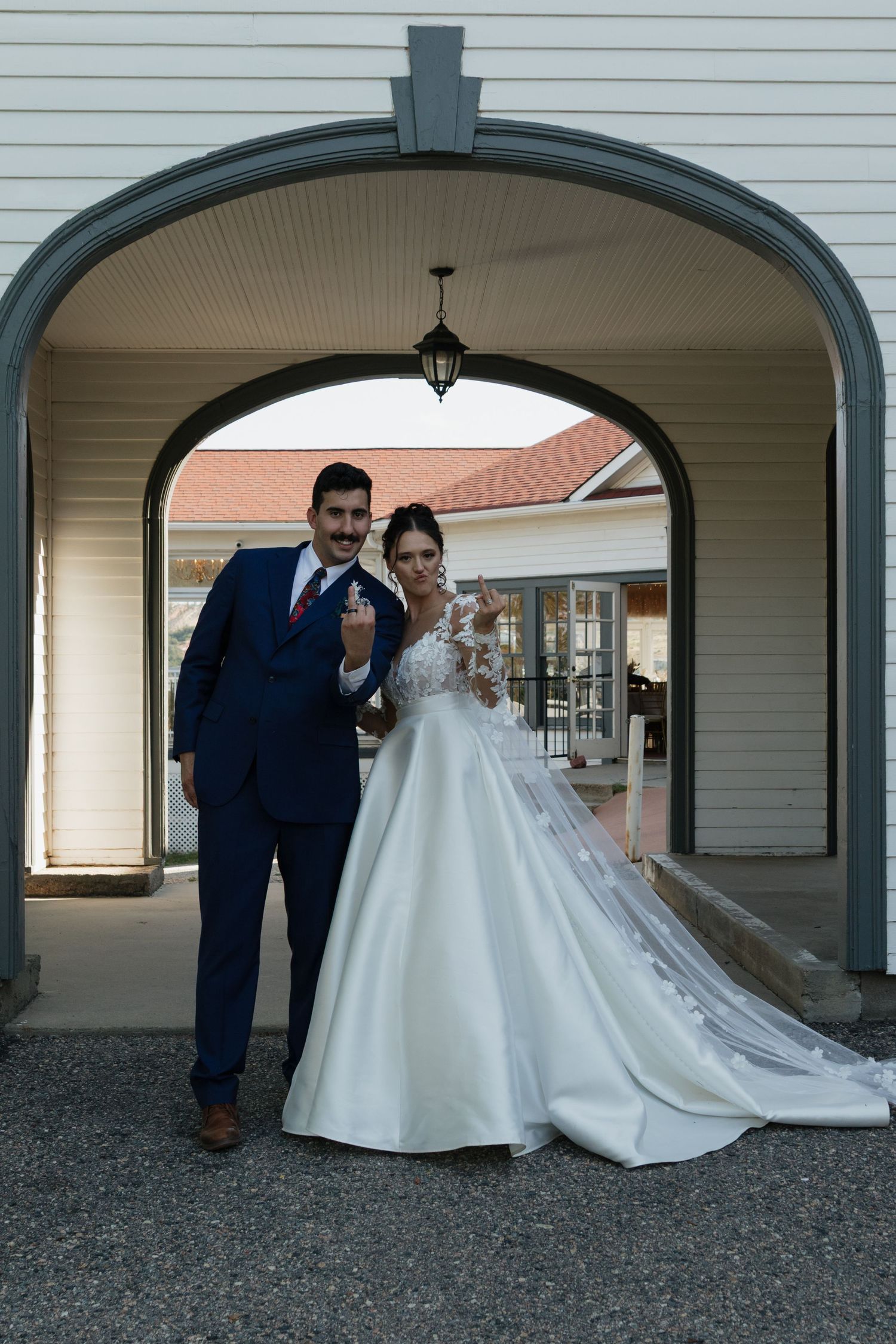 Elegant bride in flowing white gown poses under arched walkway.