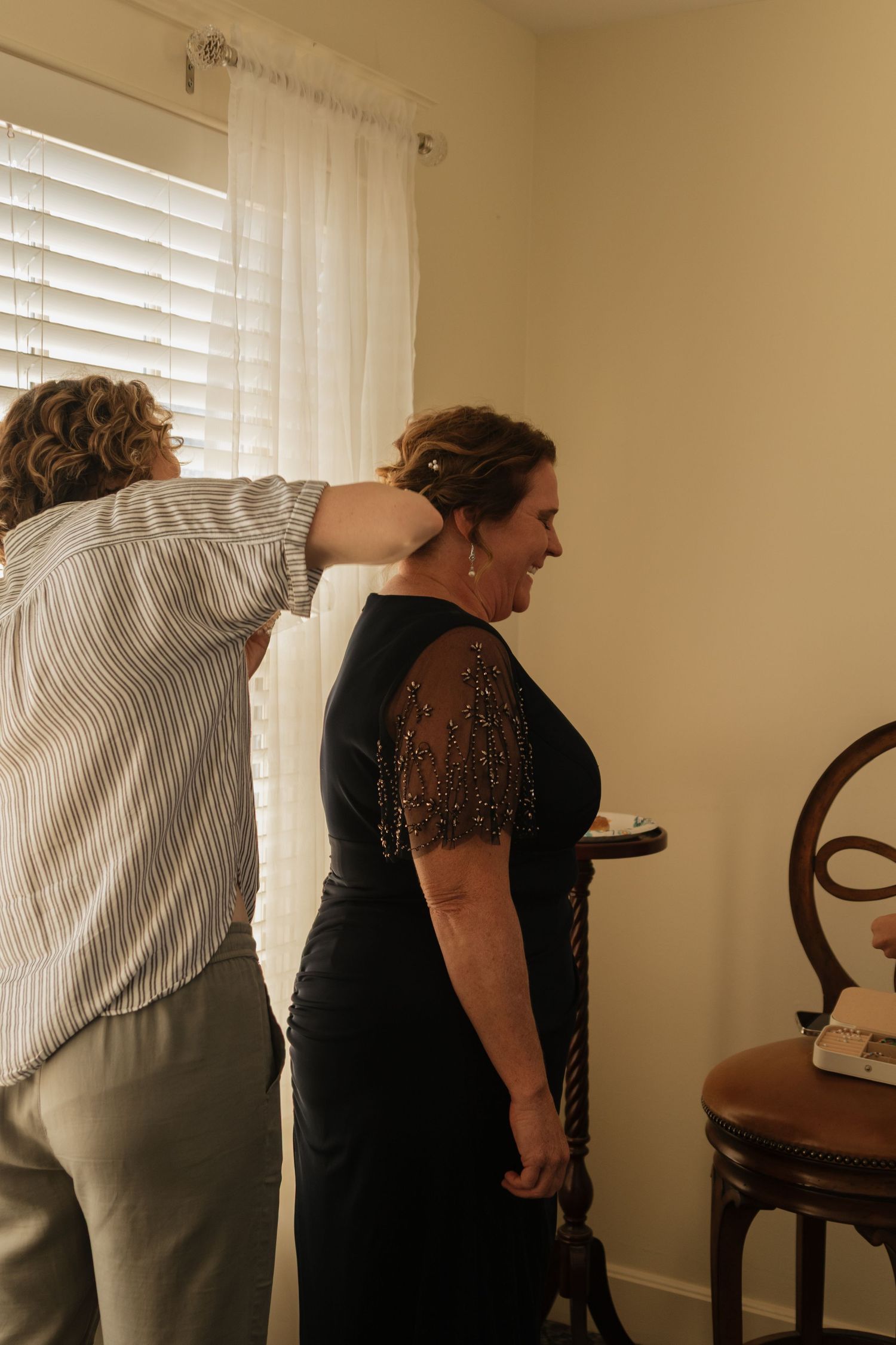 Two people in formal wear stand together in a room with beige walls and window light.