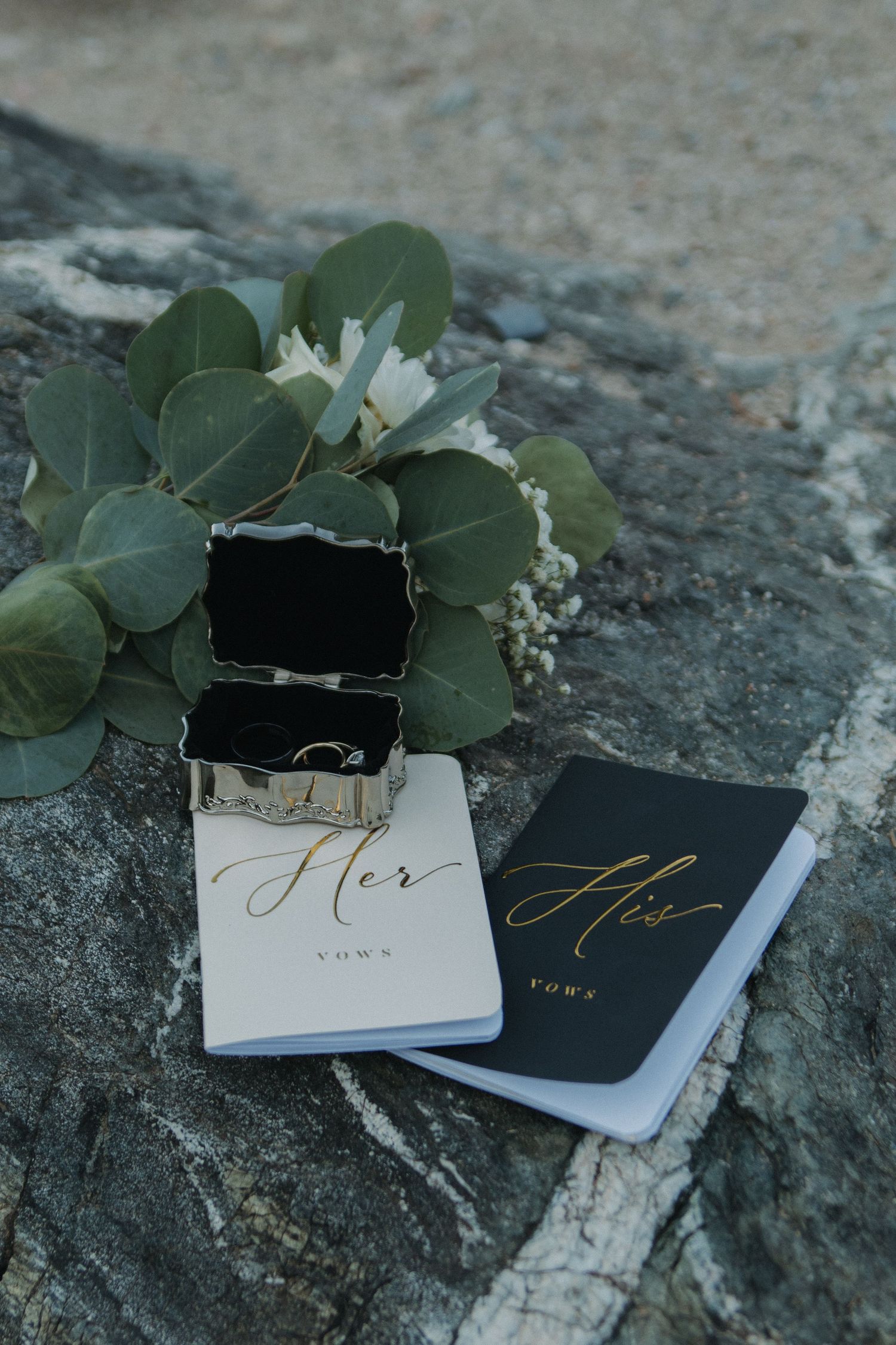 Wedding vow books in white and black with gold text lying on stone with eucalyptus leaves.
