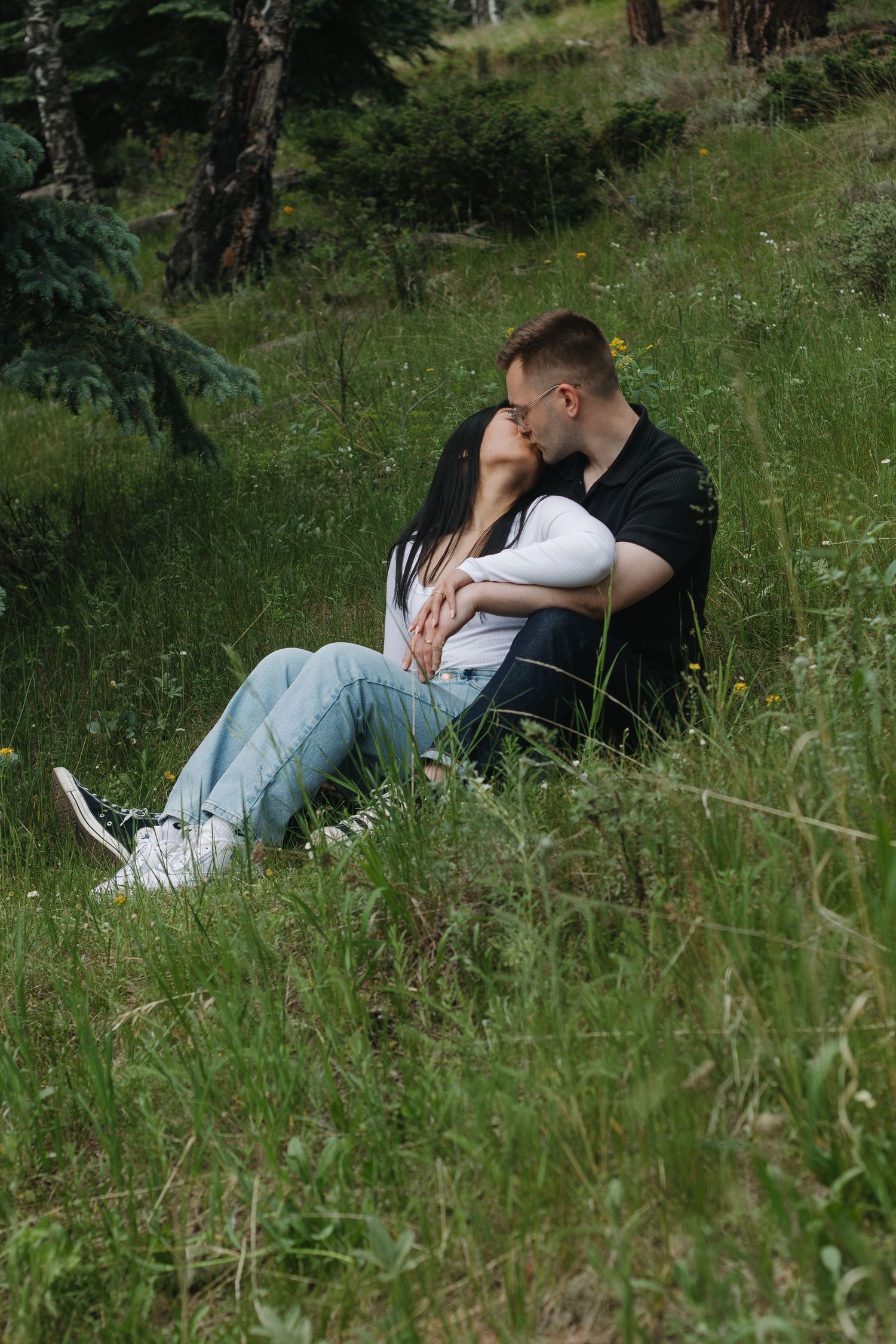 A couple shares an intimate moment while sitting in a grassy meadow surrounded by nature.