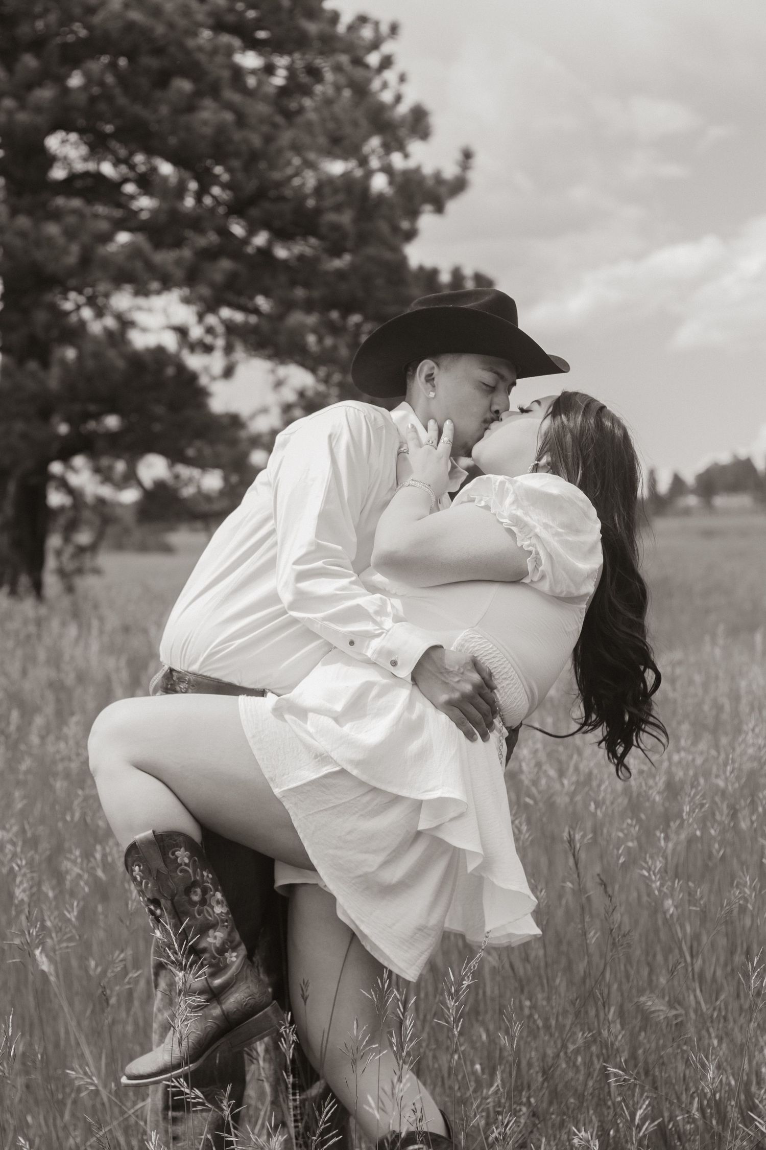 Black and white photo of romantic couple in western attire sharing passionate moment in field.