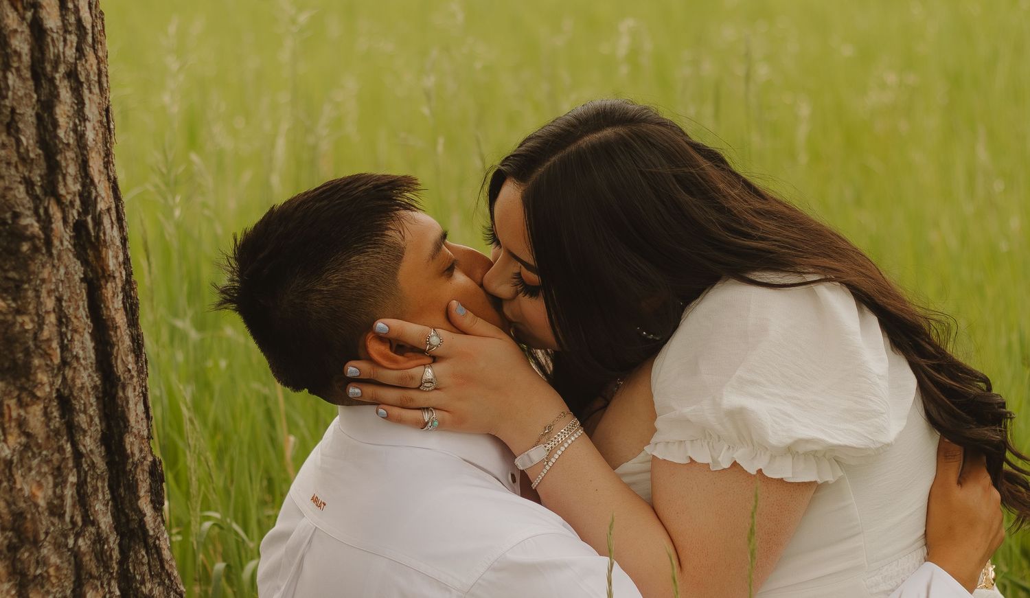 Tender moment between a couple in white clothing sharing a kiss near a tree trunk.