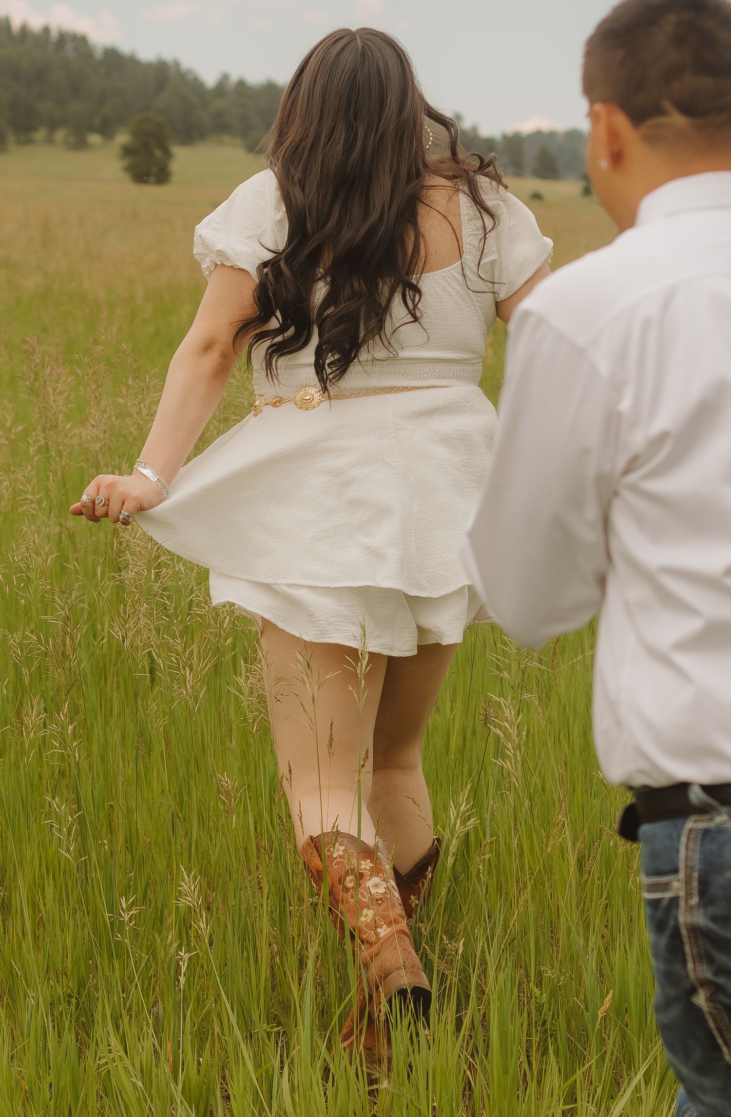 Woman in white dress and cowboy boots walking through tall grass meadow.