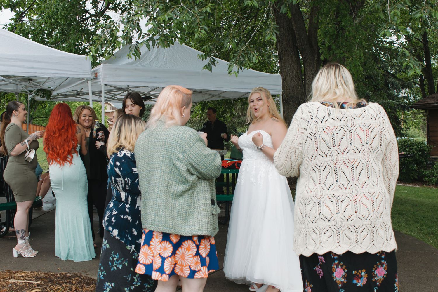 Wedding guests gather under a white tent canopy for group photos in a garden setting.