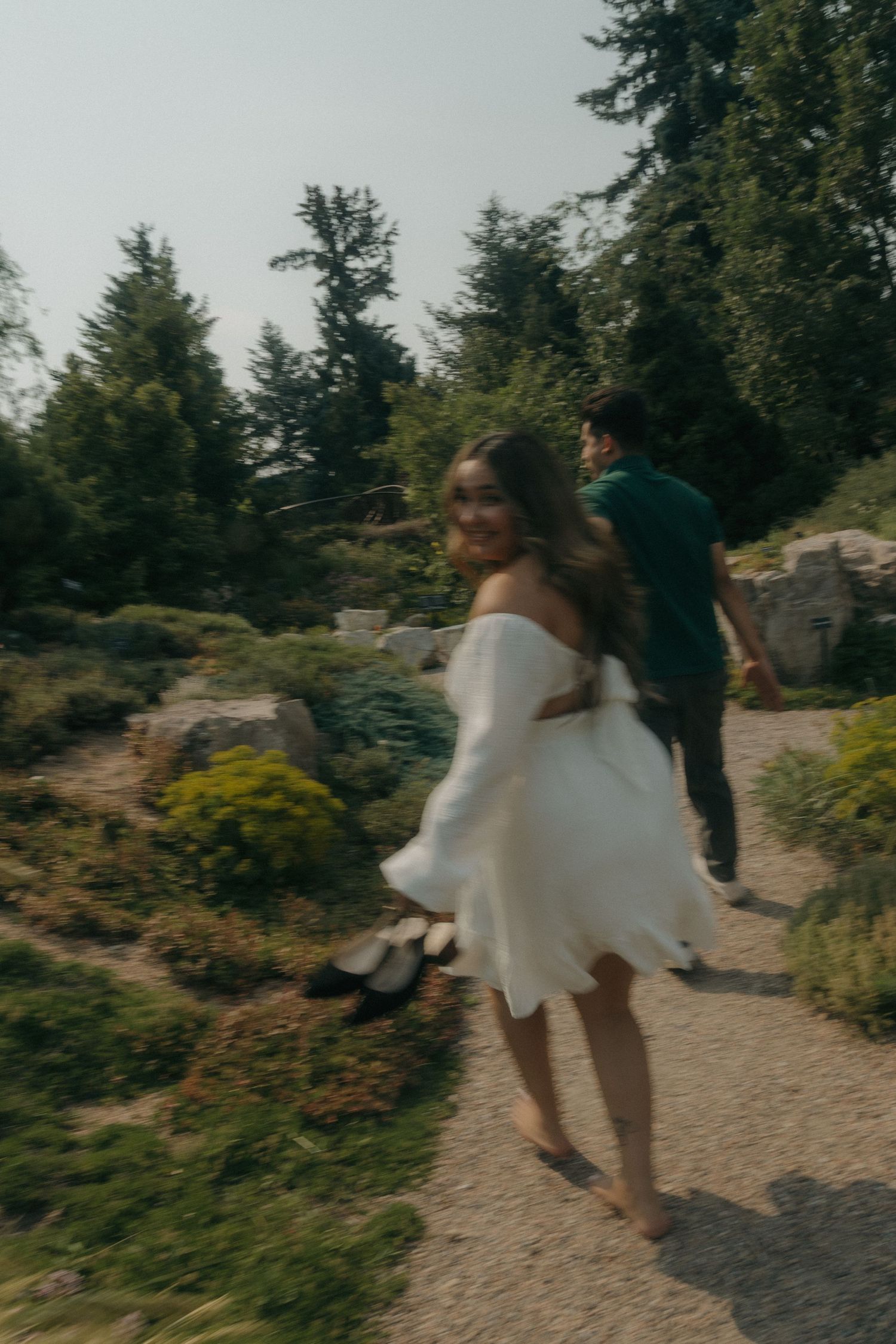 Woman in white off-shoulder dress twirling and smiling on a garden path while her partner walks ahead.