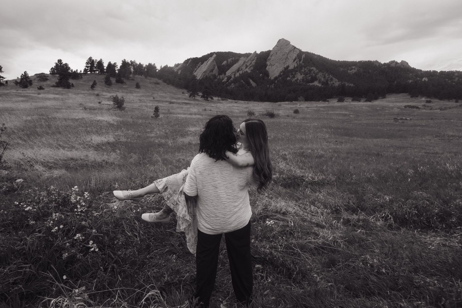 Black and white photo of a couple holding each other while gazing at dramatic flat iron mountains.