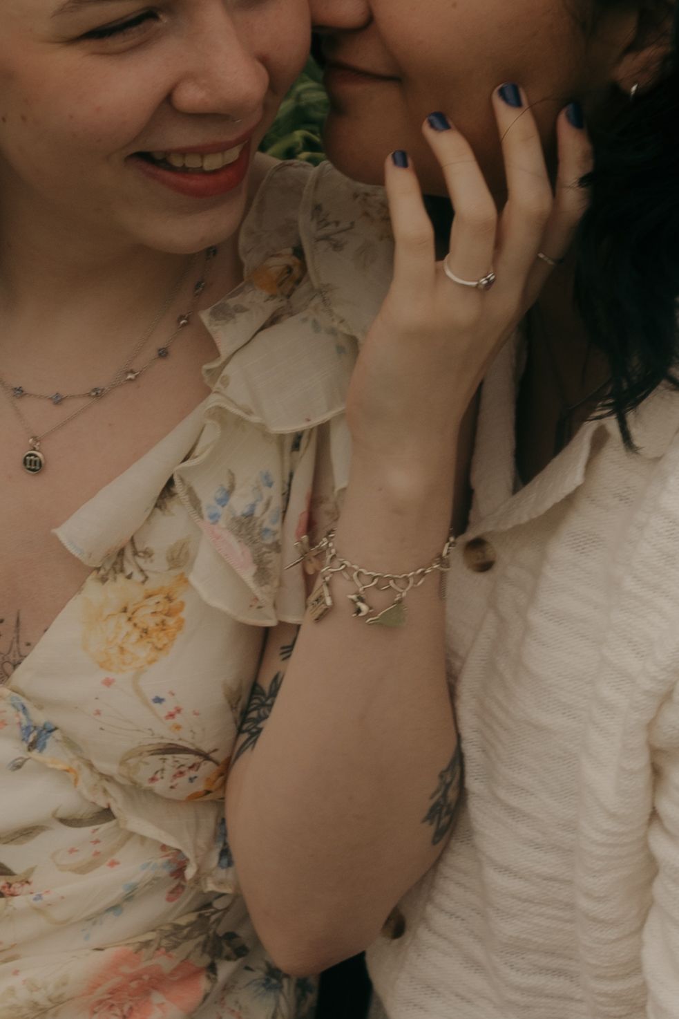 Two women sharing a tender moment, one wearing a floral dress and charm bracelet with blue nail polish.