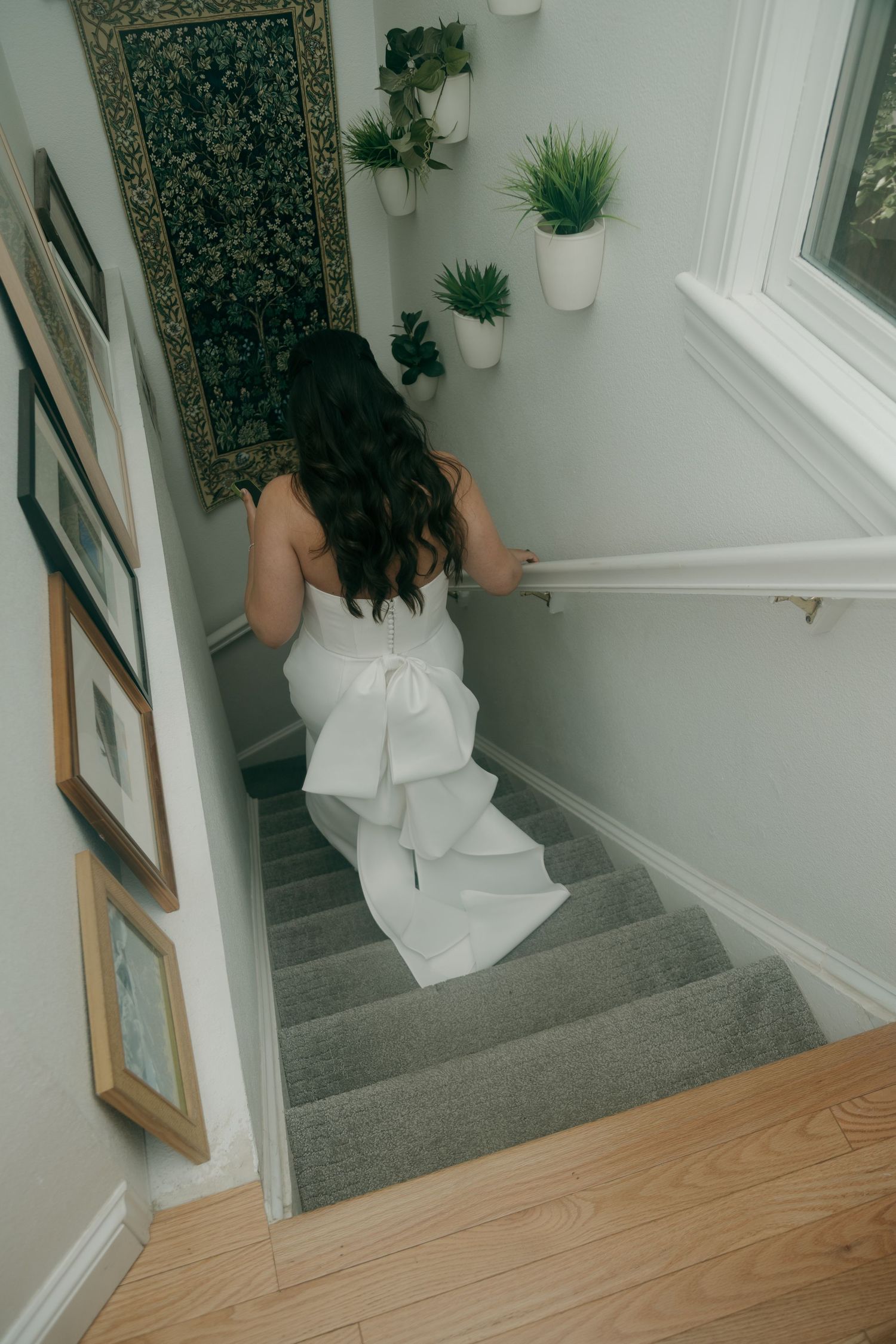 Bride in elegant white wedding gown with large bow walks up carpeted staircase.
