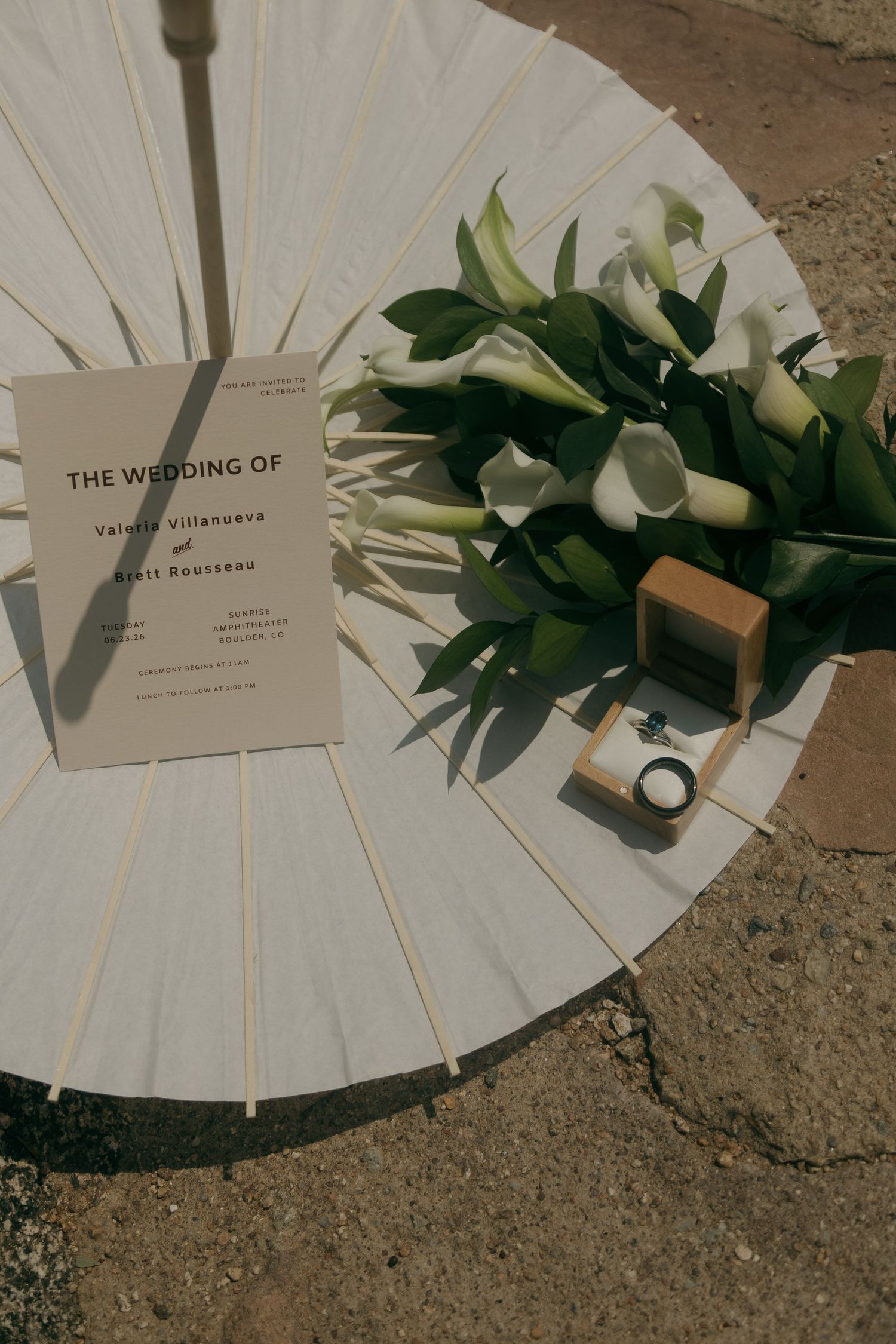 Wedding program for Valeria Villanueva and Scott Rousseau displayed on white parasol with calla lily bouquet and ring box.