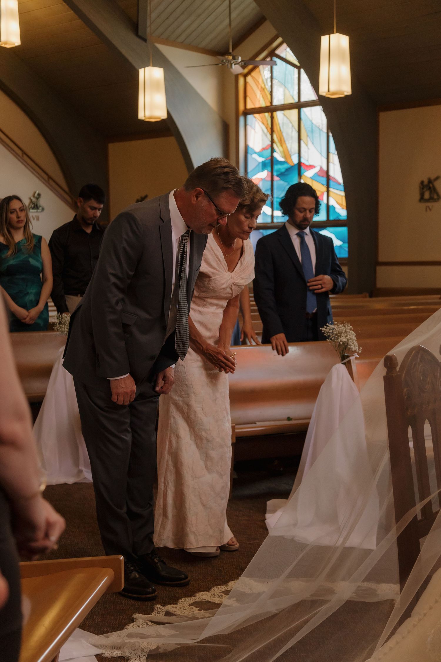 A woman in a white dress and man in a gray suit bow their heads in prayer during a church wedding ceremony.